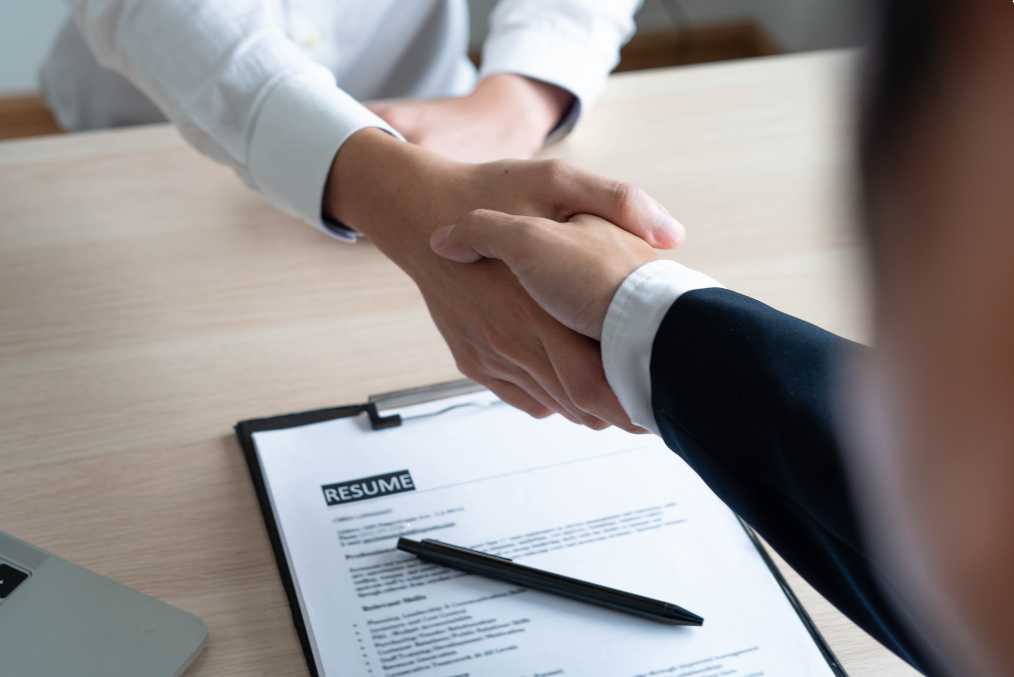 Mehrere Lebensläufe mit einer Brille darauf liegen auf einem Holztisch neben einem Laptop und einer kleinen Uhr.