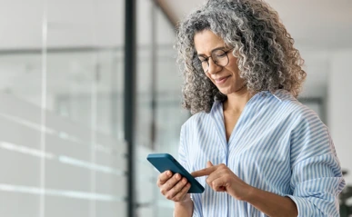 Smiling woman with curly gray hair and glasses using smartphone indoors.