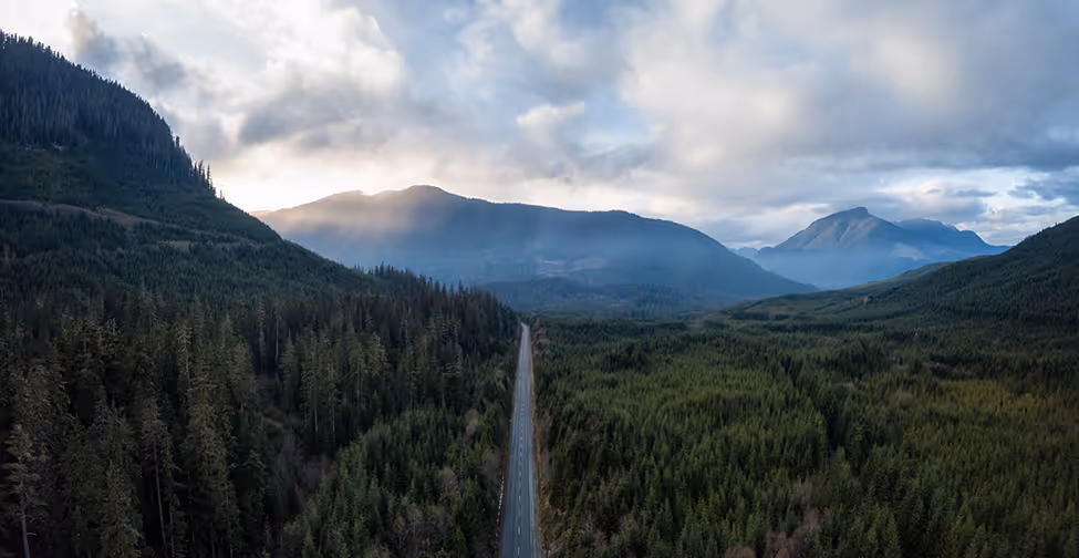 Aerial view of a road cutting through dense evergreen forest with mountains in the misty background under a partly cloudy sky.