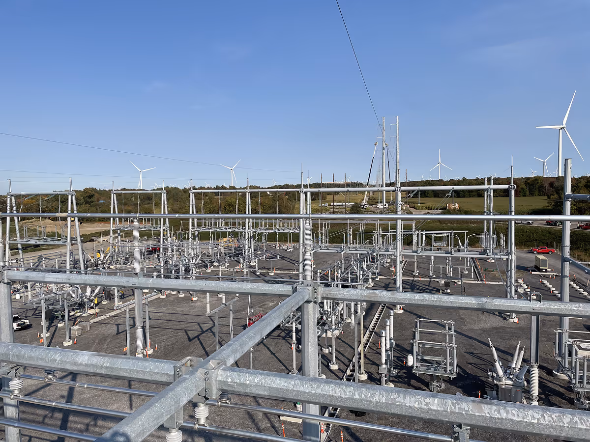 Electrical substation with metal structures and insulators under clear blue sky, with wind turbines and greenery in the background.