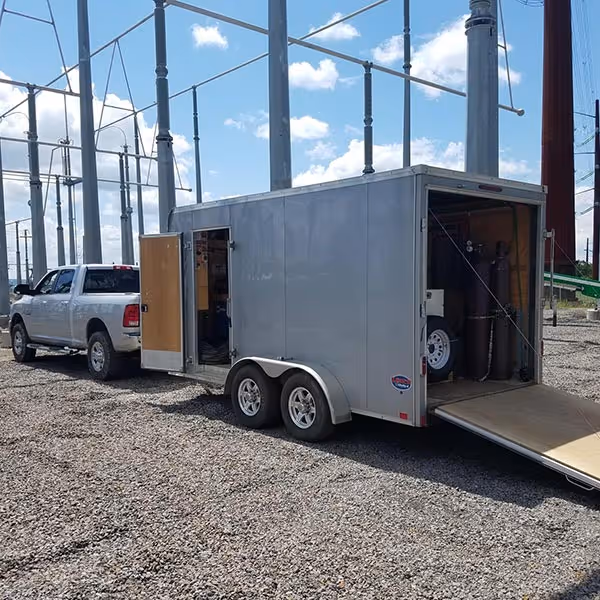 Silver pickup truck towing a silver enclosed trailer with side and rear doors open, parked on gravel under electrical poles.