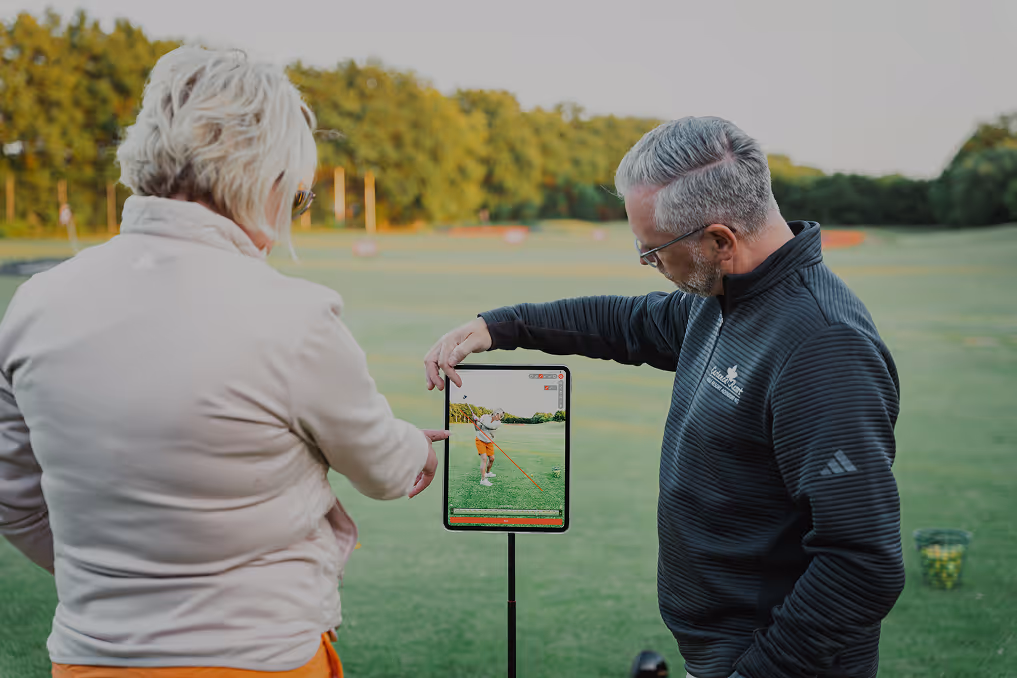 Zwei Personen auf Golfplatz schauen auf Tablet mit Video eines Golfspielers beim Abschlag.
