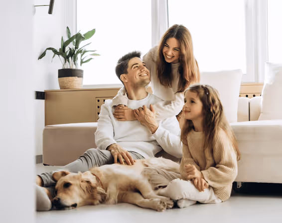 Smiling family with a man, woman, child, and a dog sitting together on a living room floor.