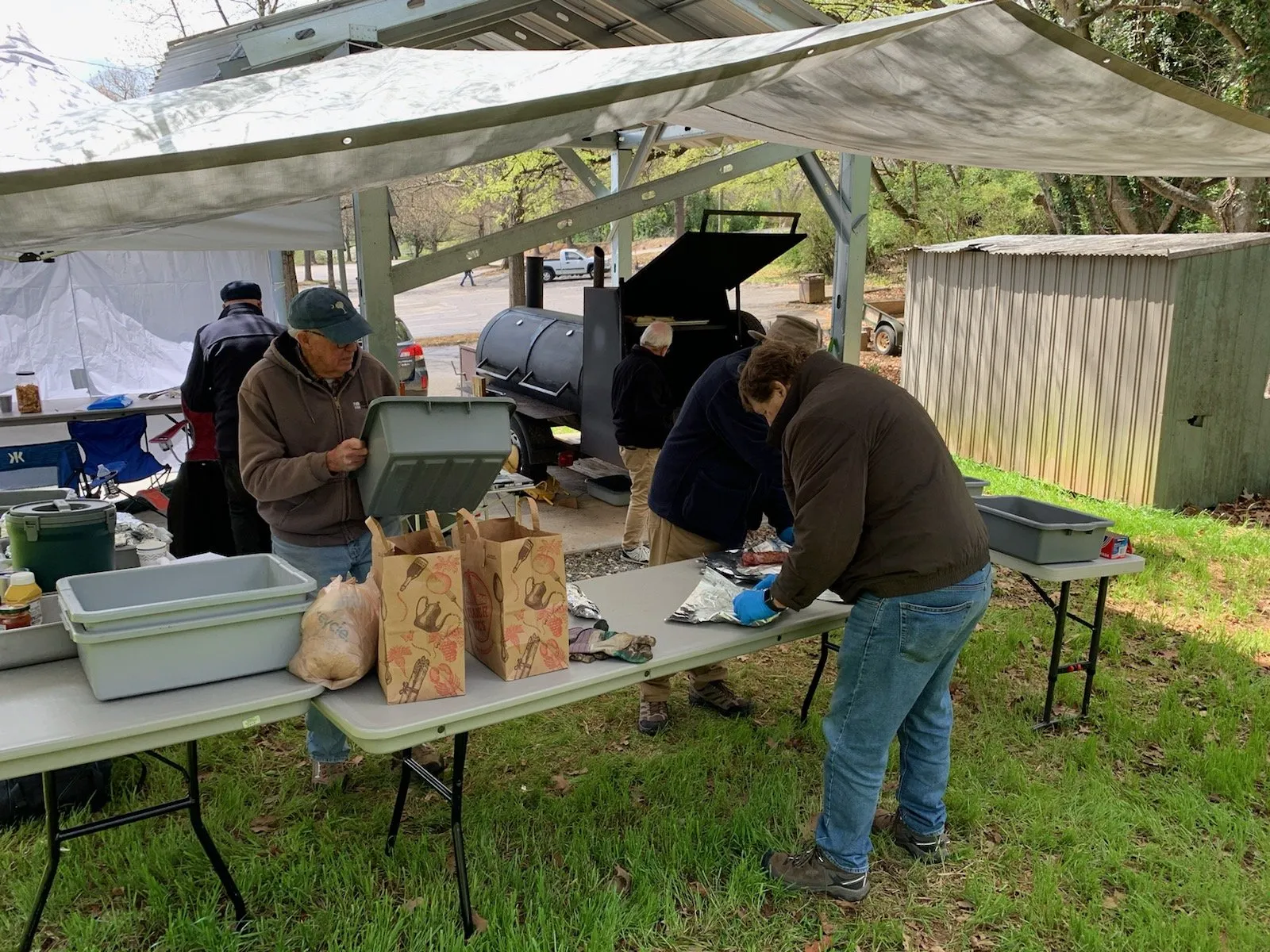 Several people prepare and wrap food outdoors under a canopy near a large barbecue smoker and folding tables with bags and containers.