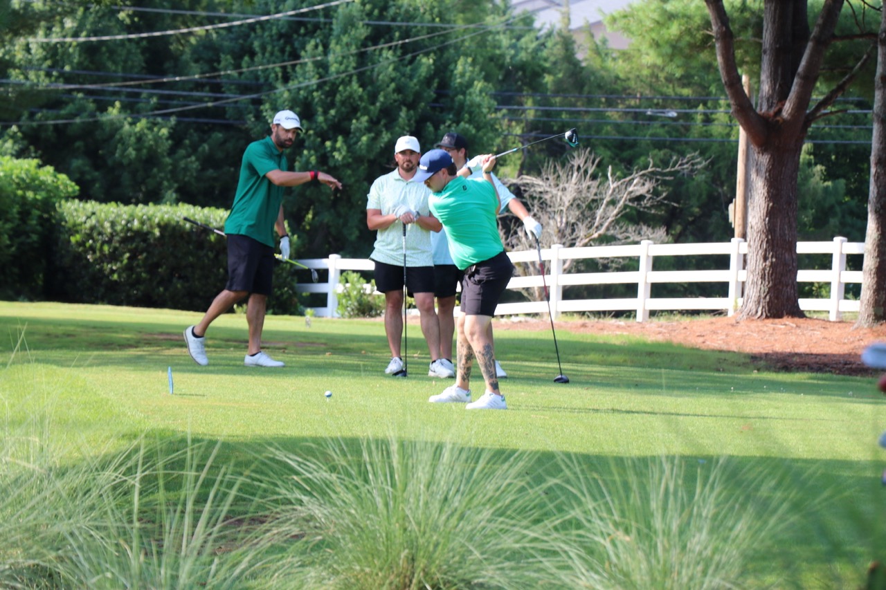 Four men on a golf course, one swinging a club while the others watch and point.
