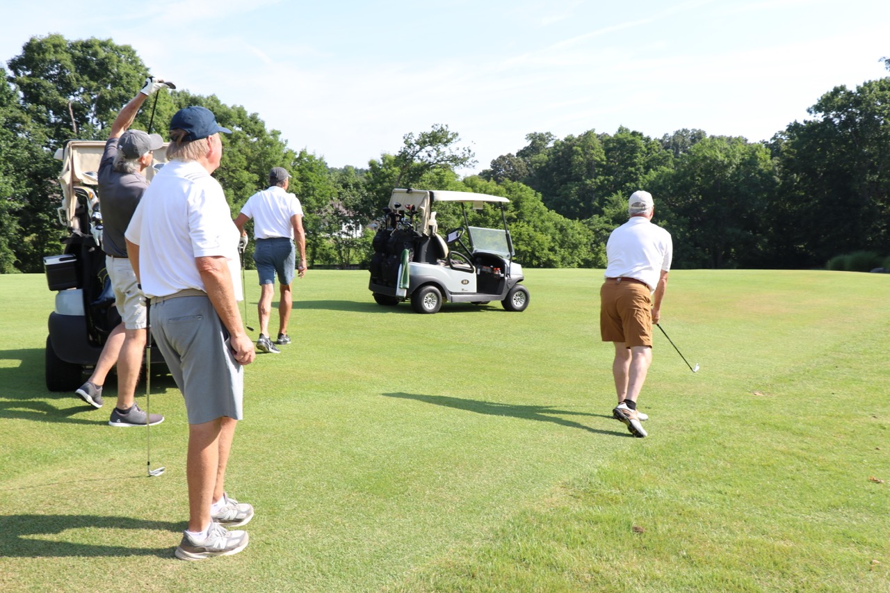 Four men on a golf course, one preparing to hit a golf ball while others watch near two golf carts.