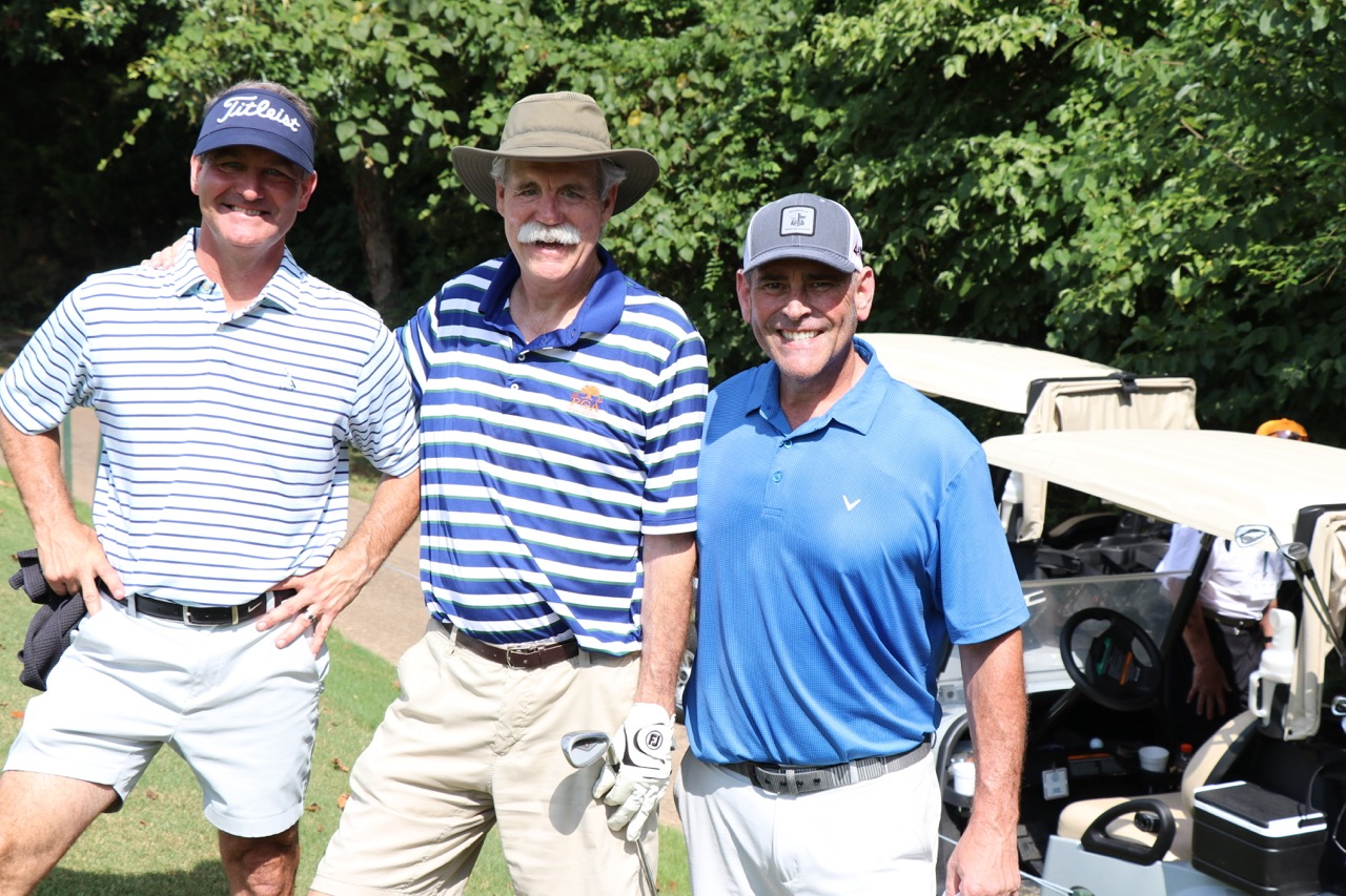Three men smiling and posing together on a golf course, two wearing striped shirts and one in a blue polo, with golf carts and trees in the background.