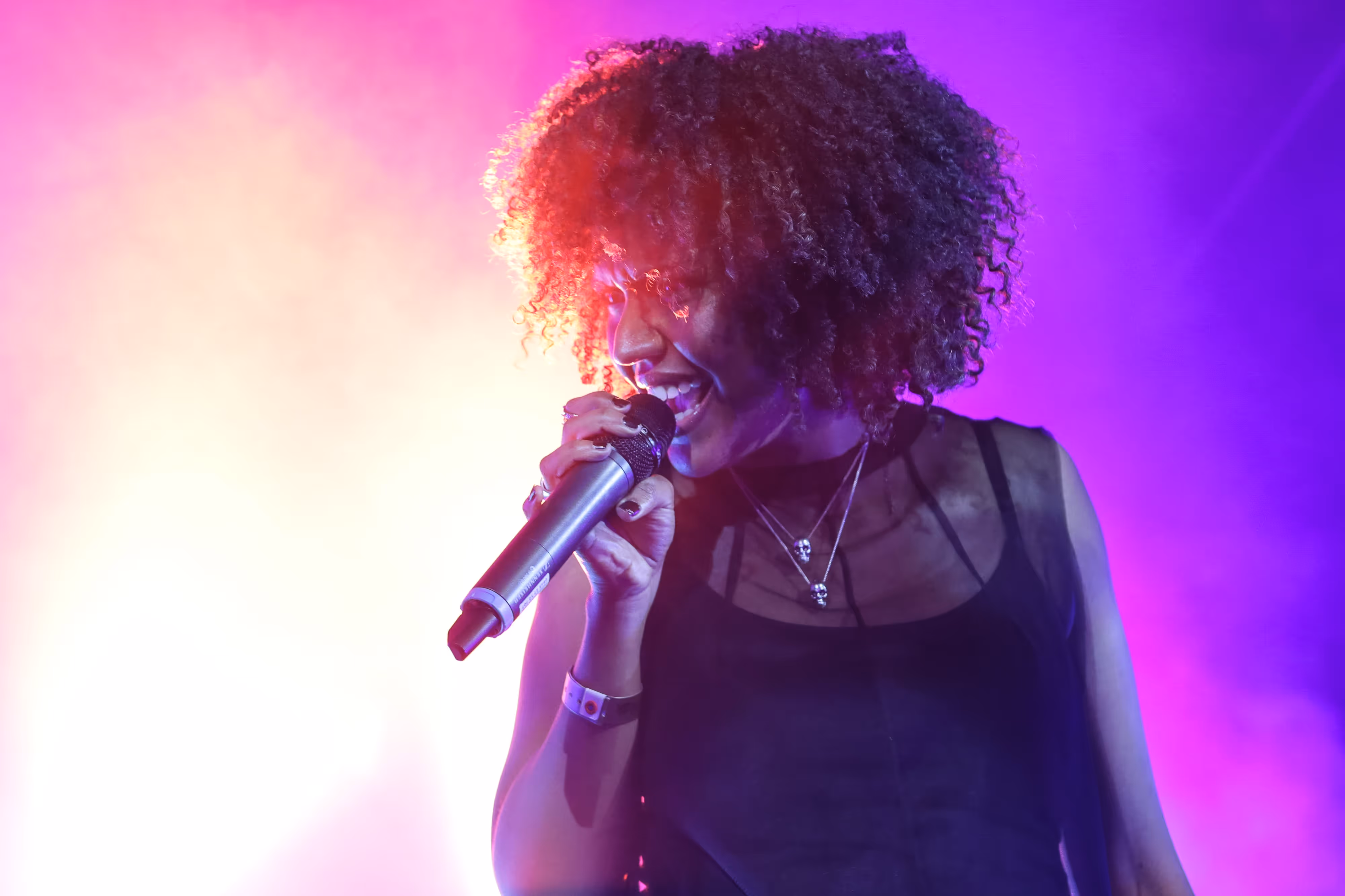 Capturing a live moment: a curly-haired singer performs into a mic, vibrant pink and purple stage lights illuminating the scene.