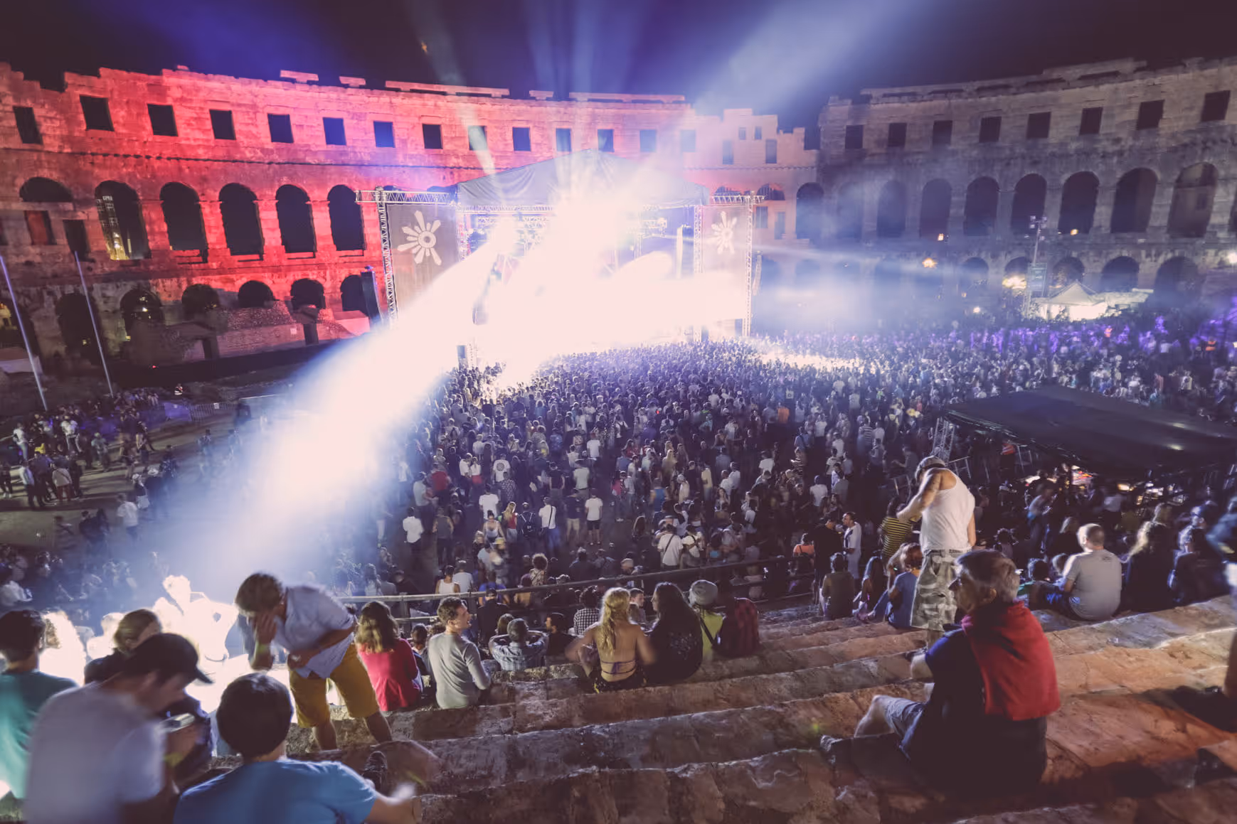 Shot of a packed audience at night in an ancient amphitheatre, vivid stage lights and colourful walls highlighting the live music energy.