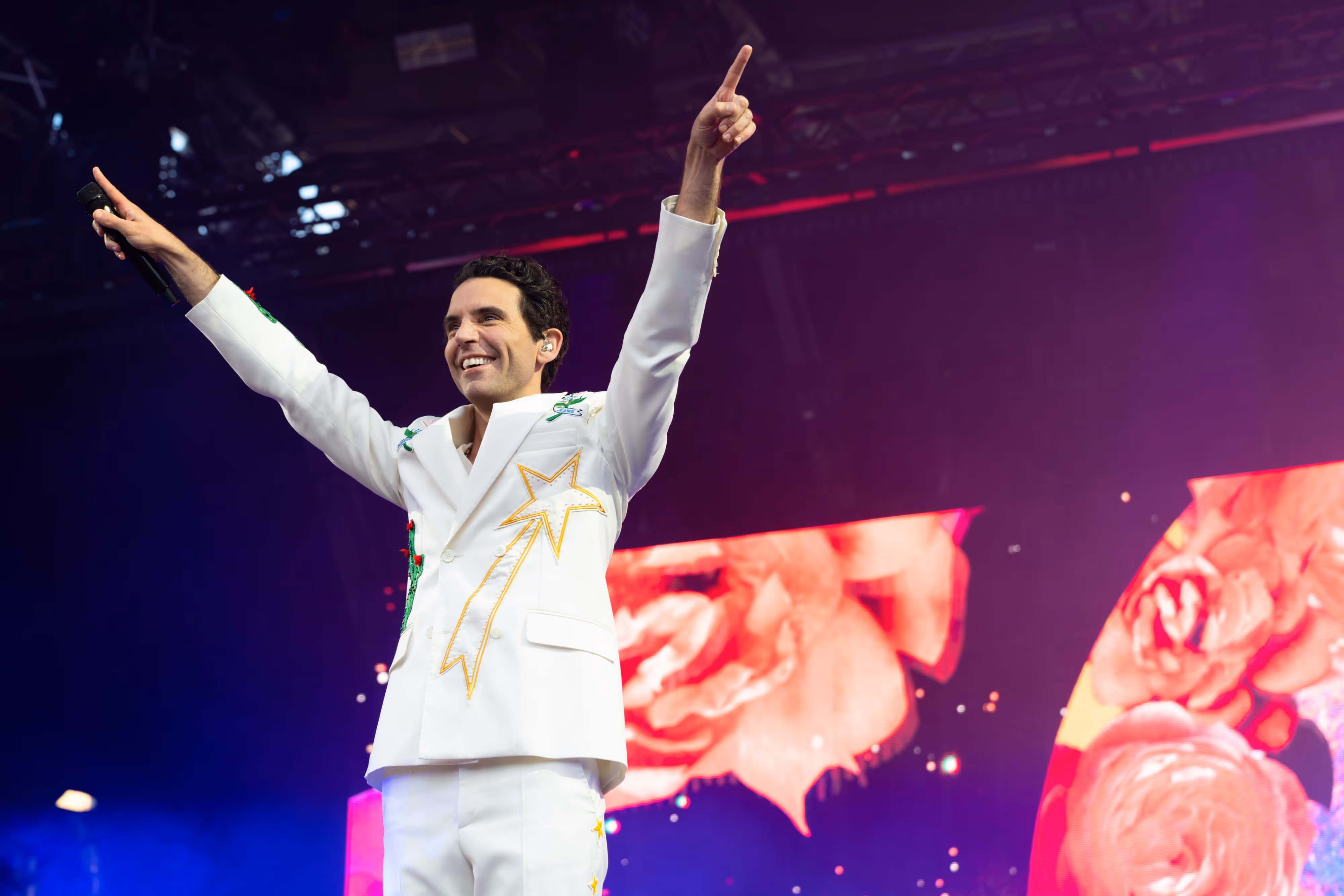 Mika performs in a white suit with vivid designs lifts both arms on a vibrant, flower-themed stage.