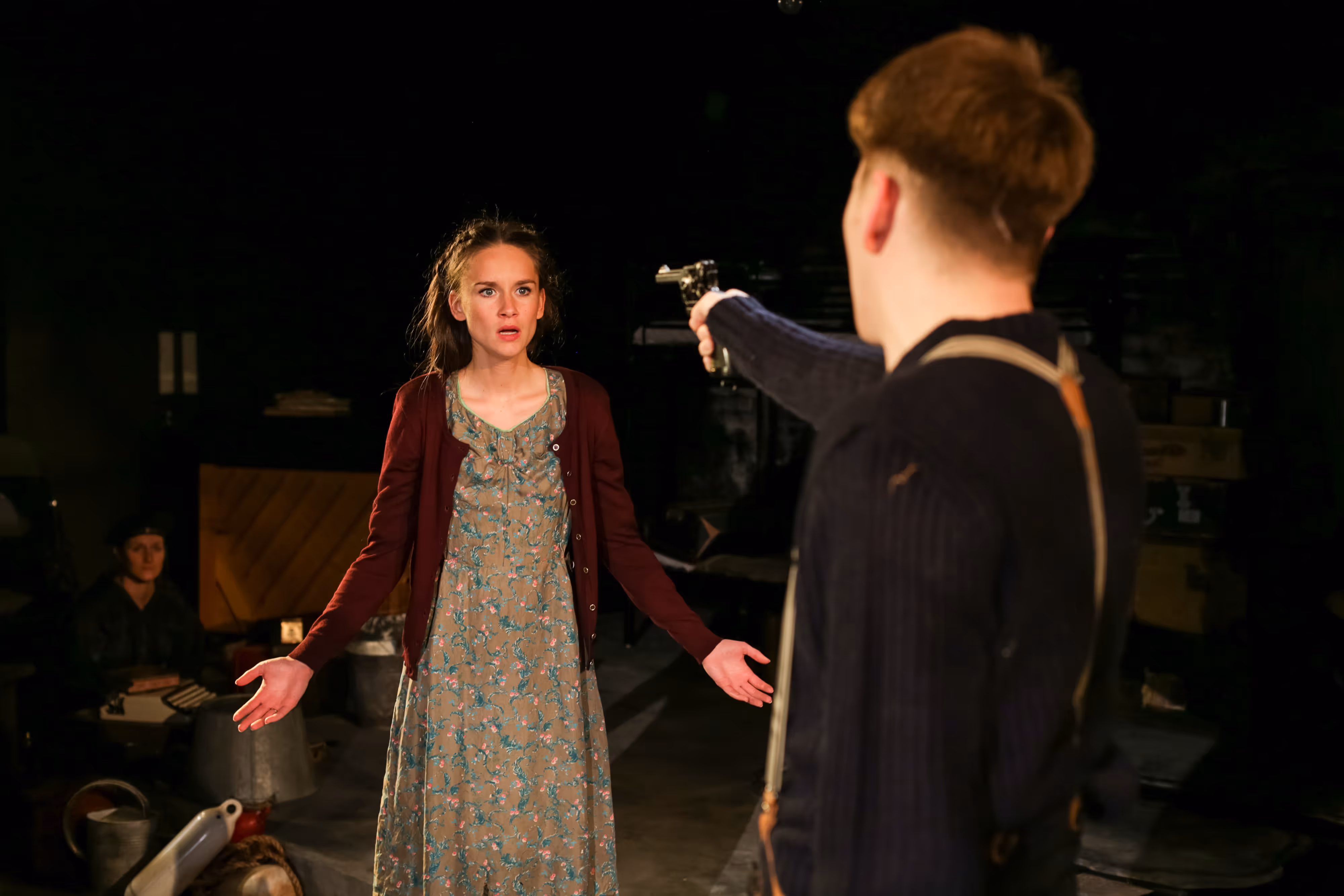 A stage actor aims a prop gun at an actress with outstretched arms in moody theatre lighting, both showing dramatic tension.