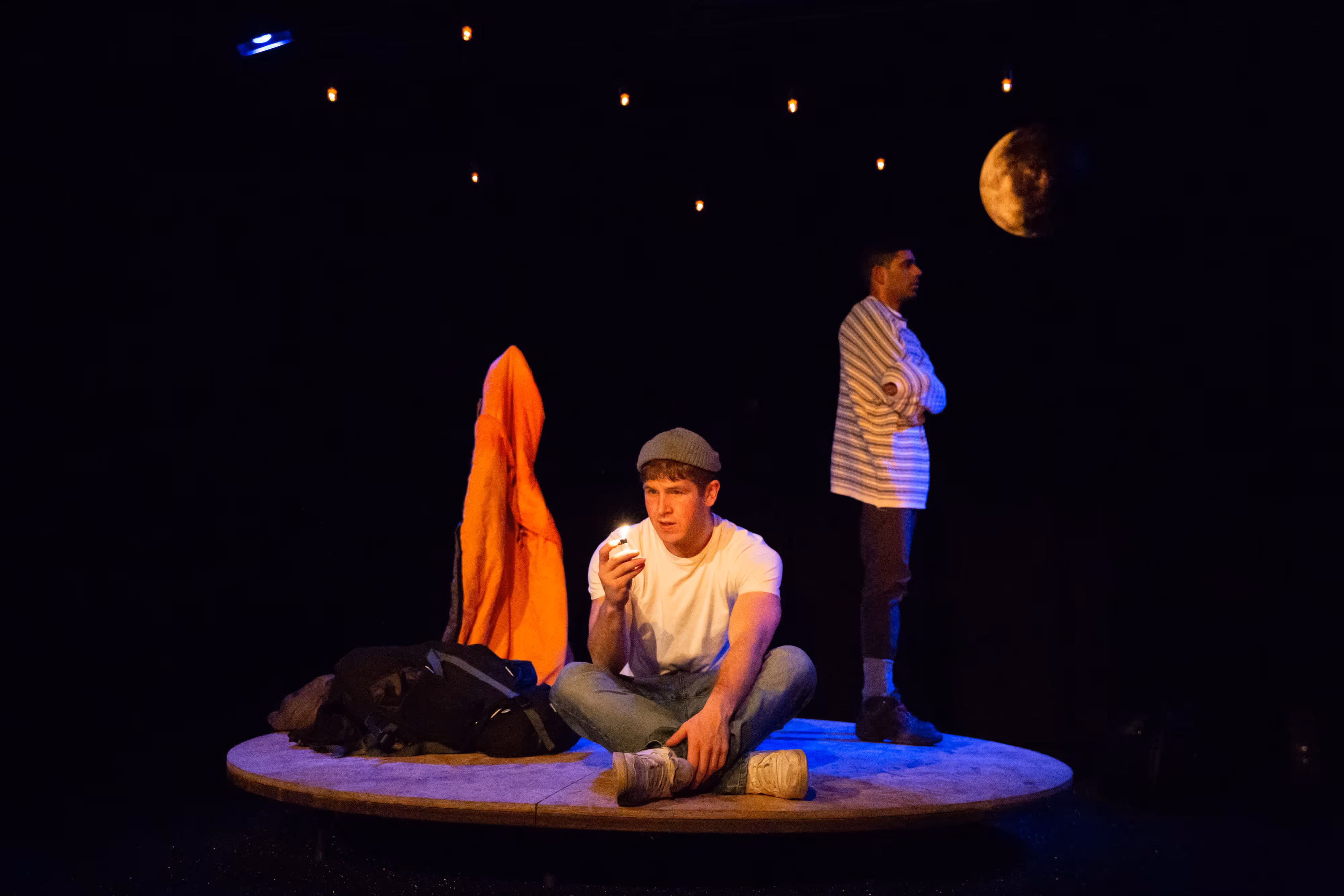 Photographed in low stage light: one actor seated, striking a match; another stands backlit by fairy lights and a crescent moon.
