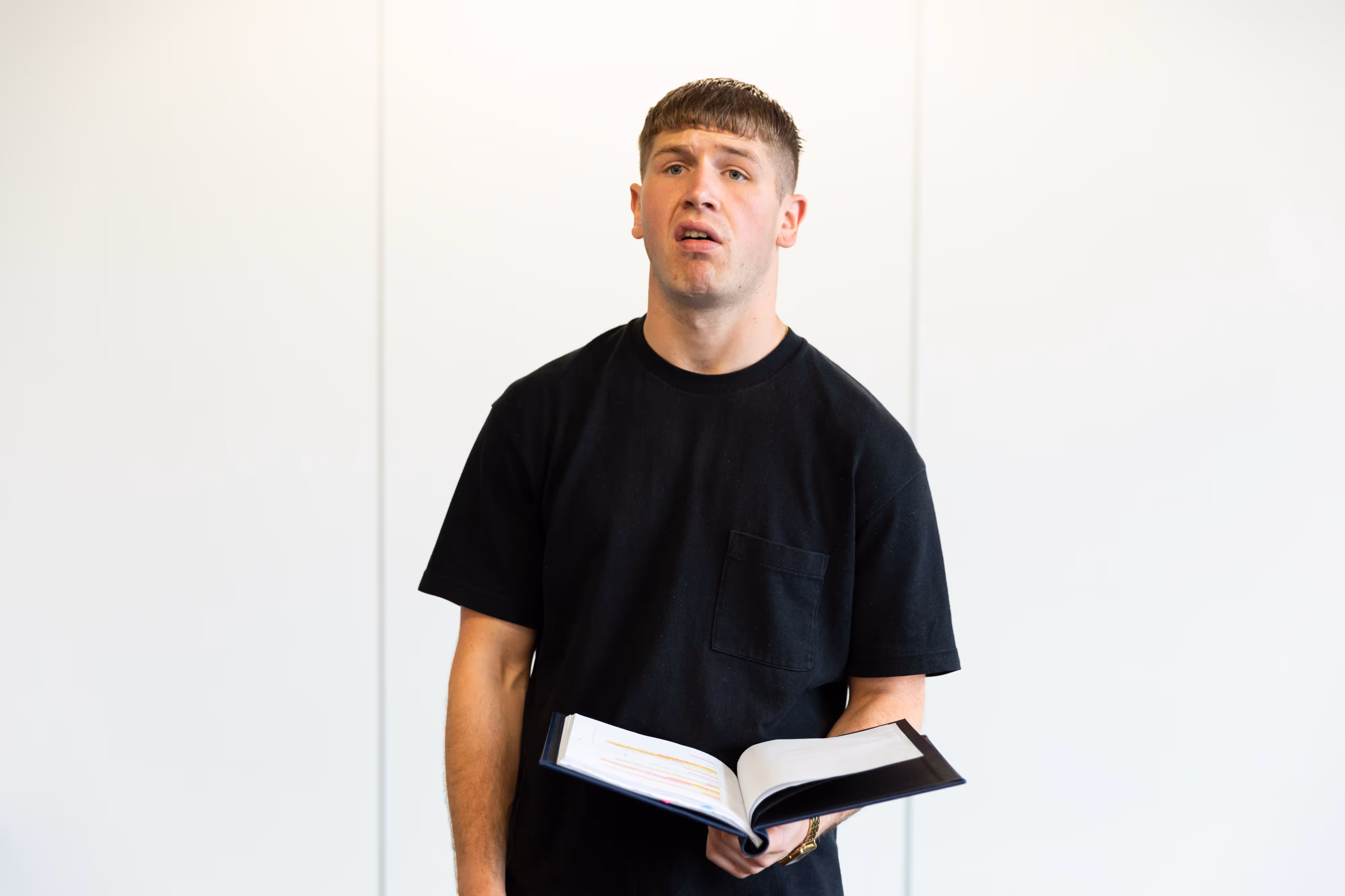 On stage, a young man in a black shirt holds an open book, gazing upwards pensively as theatre lights highlight his expression.