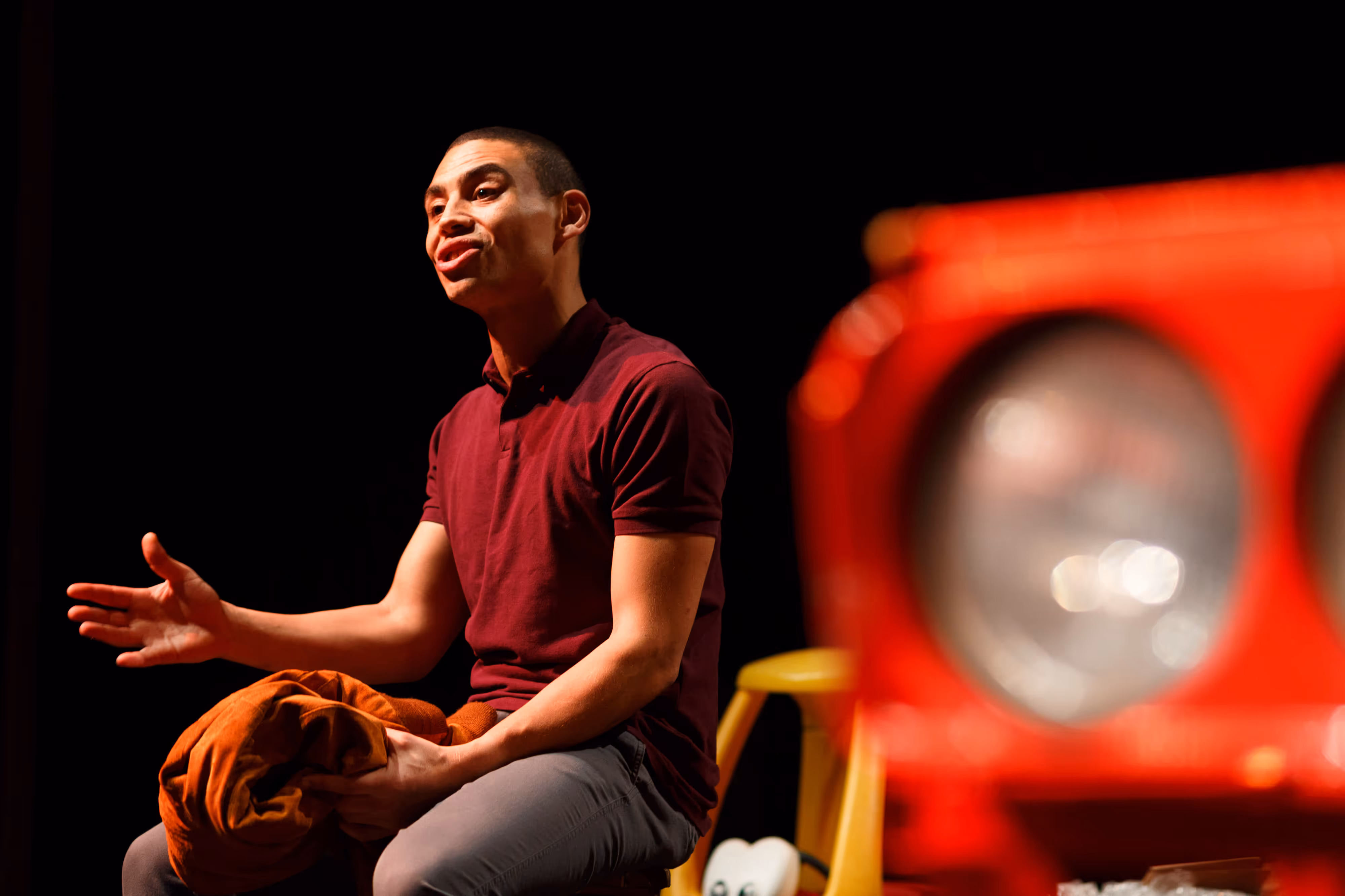 Photographed from the wings, a performer sits centre stage, gesturing mid-speech, with a blurred red prop in the foreground.