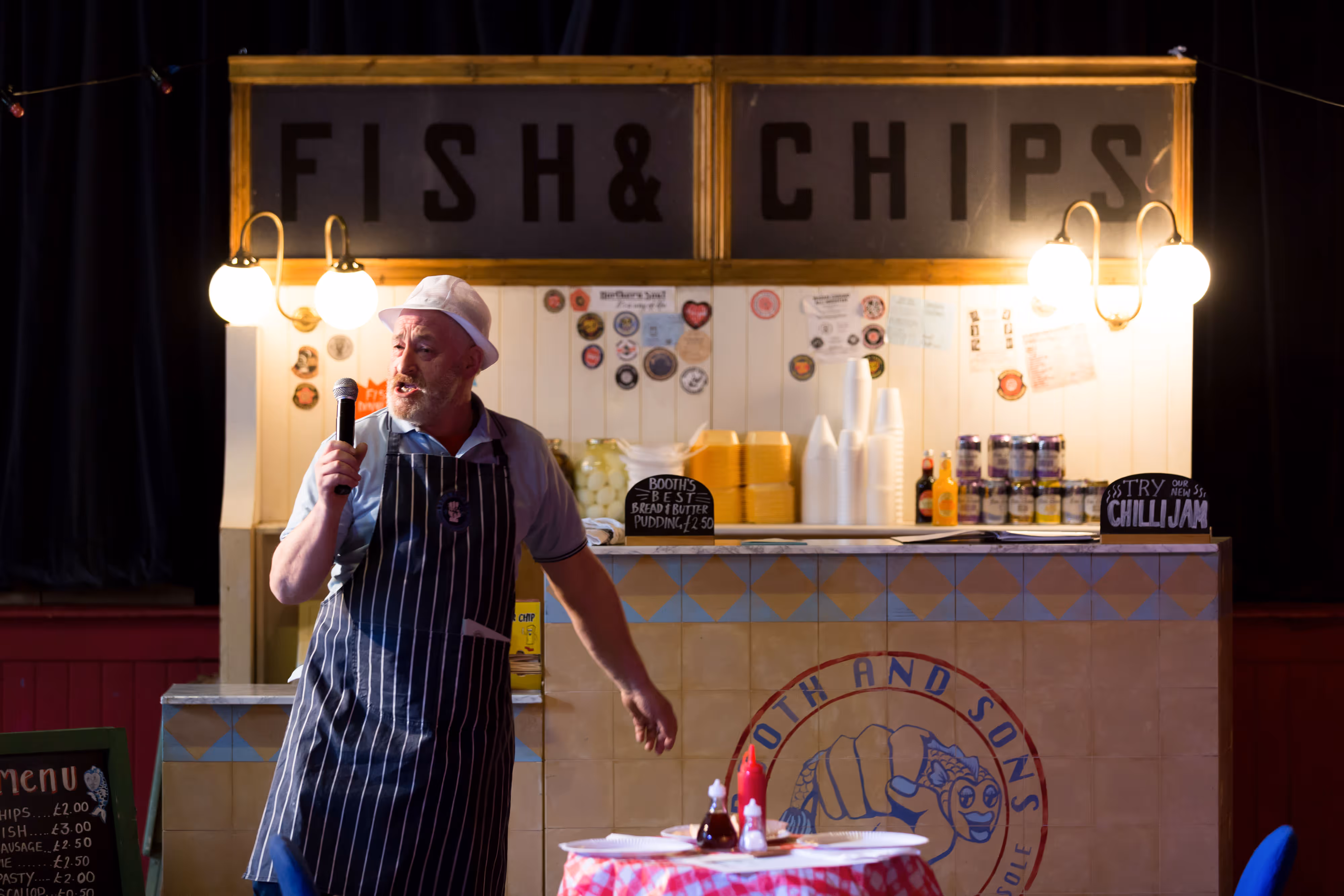 A performer in apron and hat stands at a fish and chip counter, singing animatedly into a microphone under stage lights.