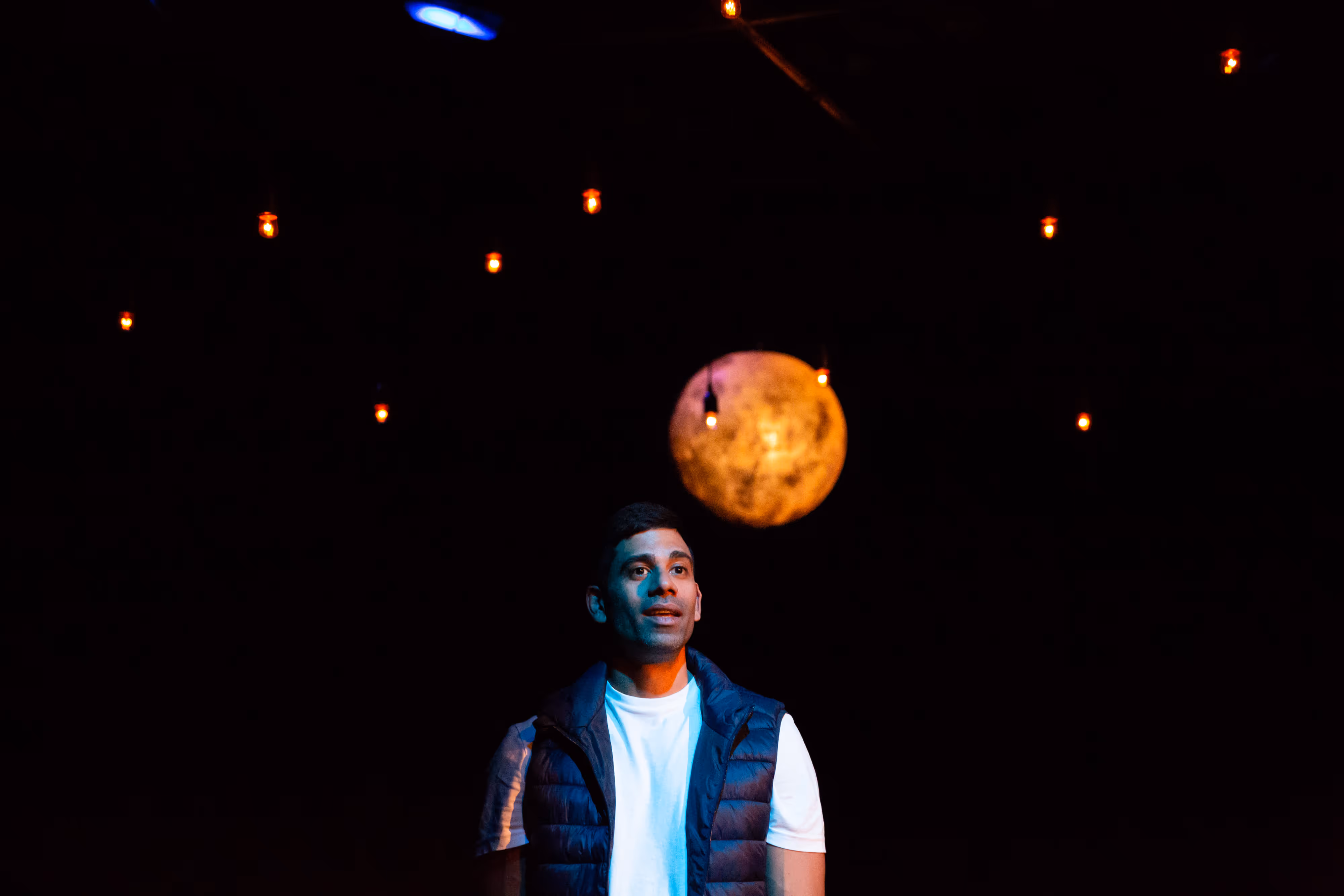 Under moody theatre lighting, a man poses beneath fairy lights and a luminous moon-like orb against a shadowy stage backdrop.