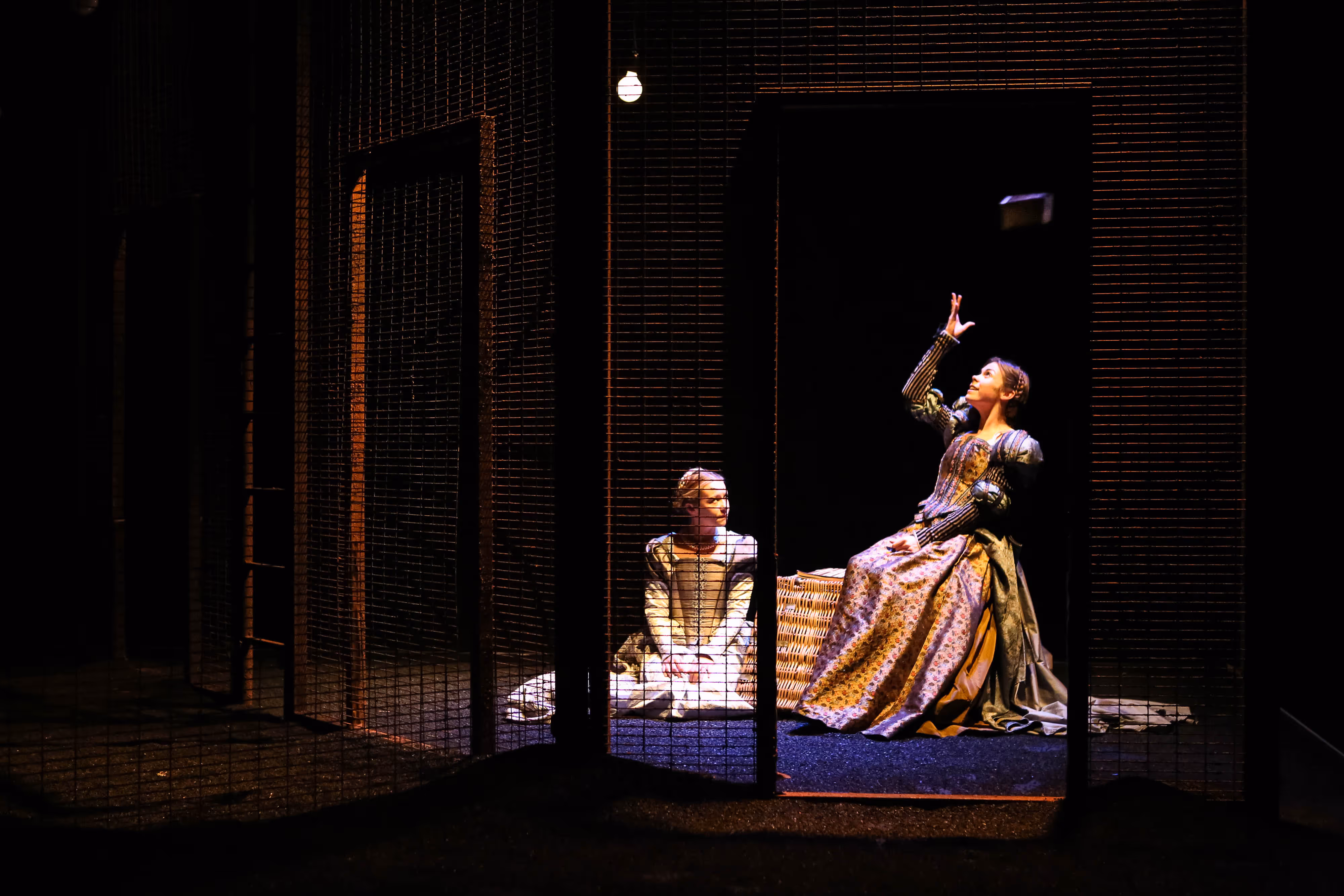 Captured onstage: two actresses in period dress, one mid-throw with a prop, bathed in moody theatrical lighting.