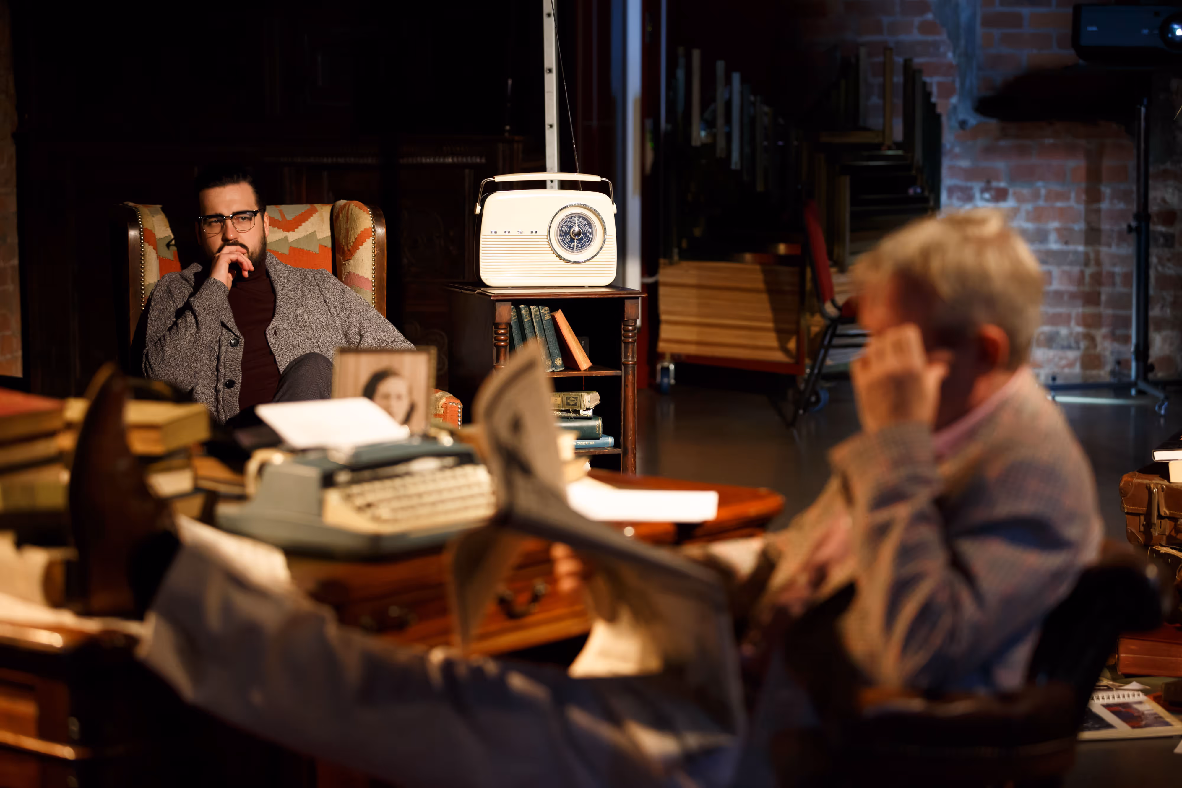 On stage, two actors in a warmly lit, book-lined set—one reading at a desk, the other listening by a vintage wireless between them.