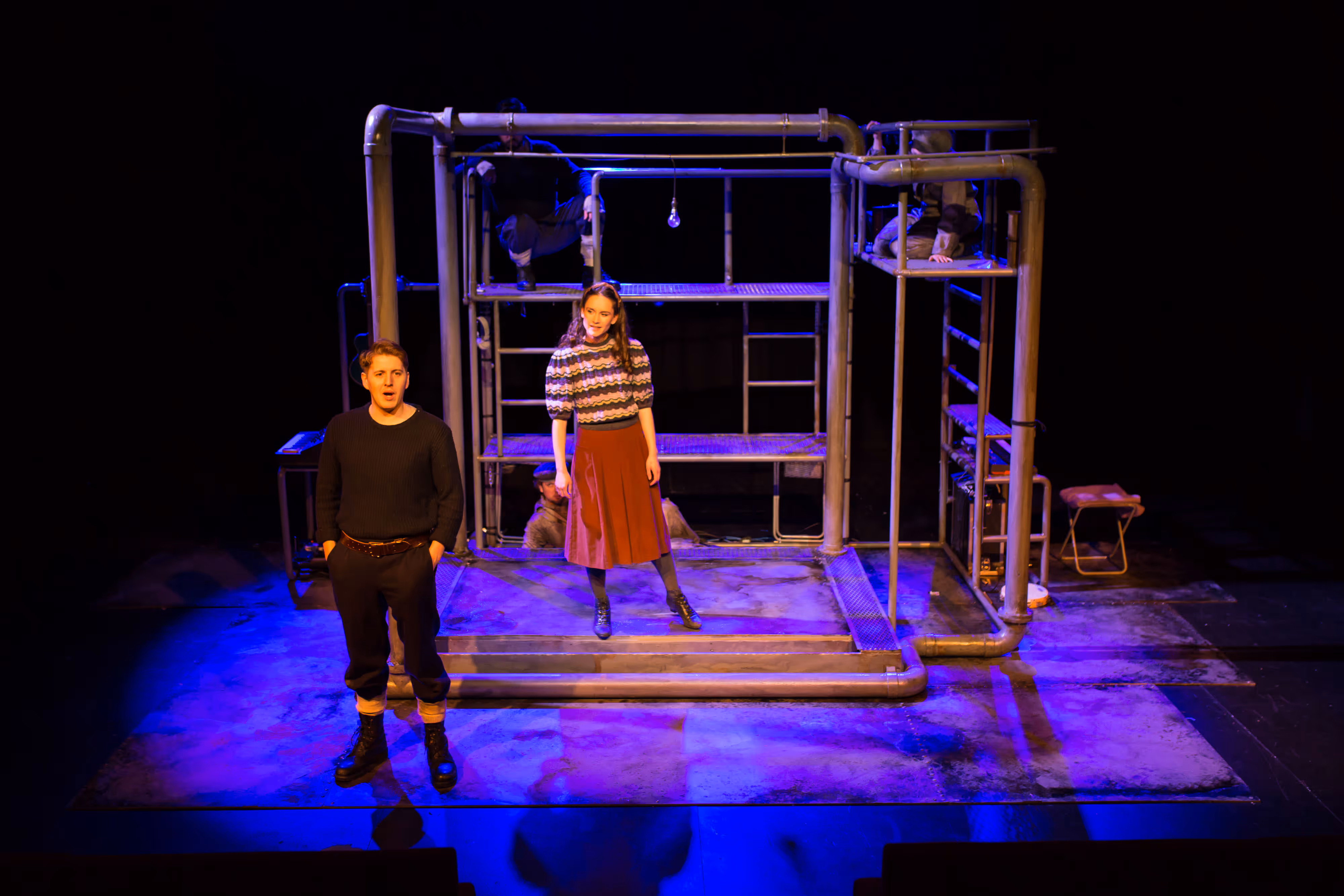Photograph of two actors onstage, framed by metal pipes and platforms, illuminated by striking blue and yellow stage lighting.
