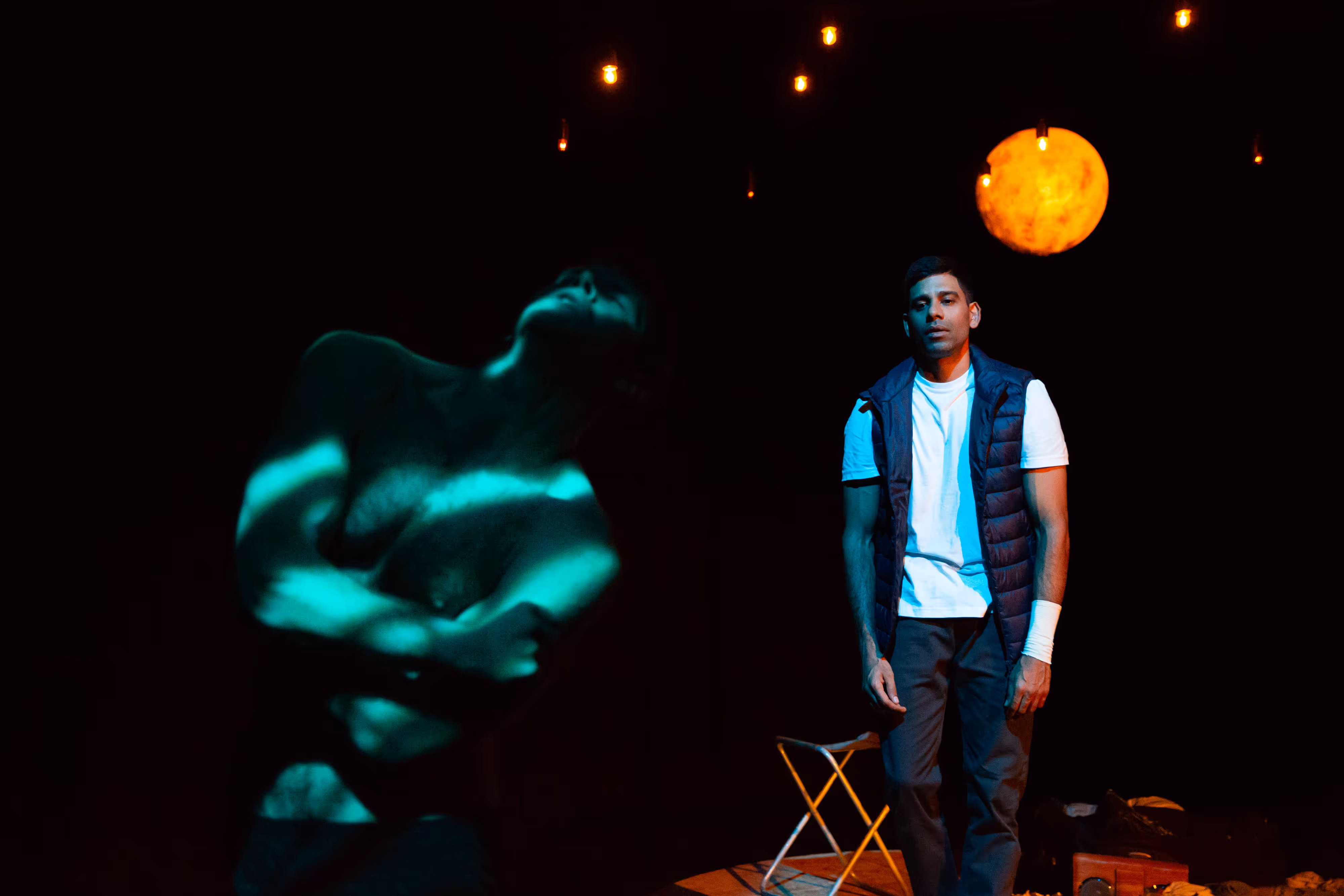 Onstage under theatrical lights, an actor stands beside a projected photo of a bare-chested figure with arms crossed.