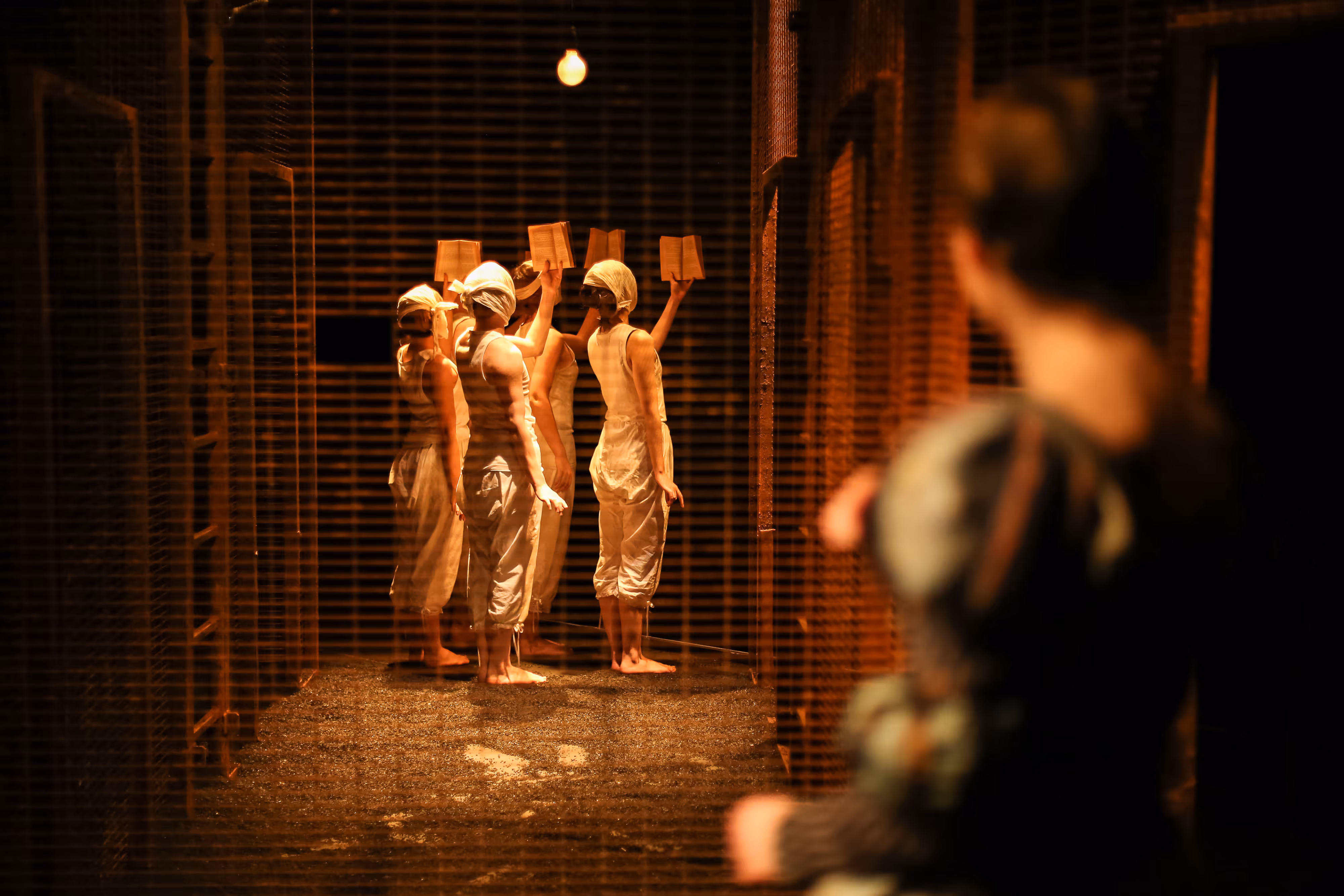 From the wings, three actors in white costumes and head coverings face upstage, books in hand, as another observes in front.