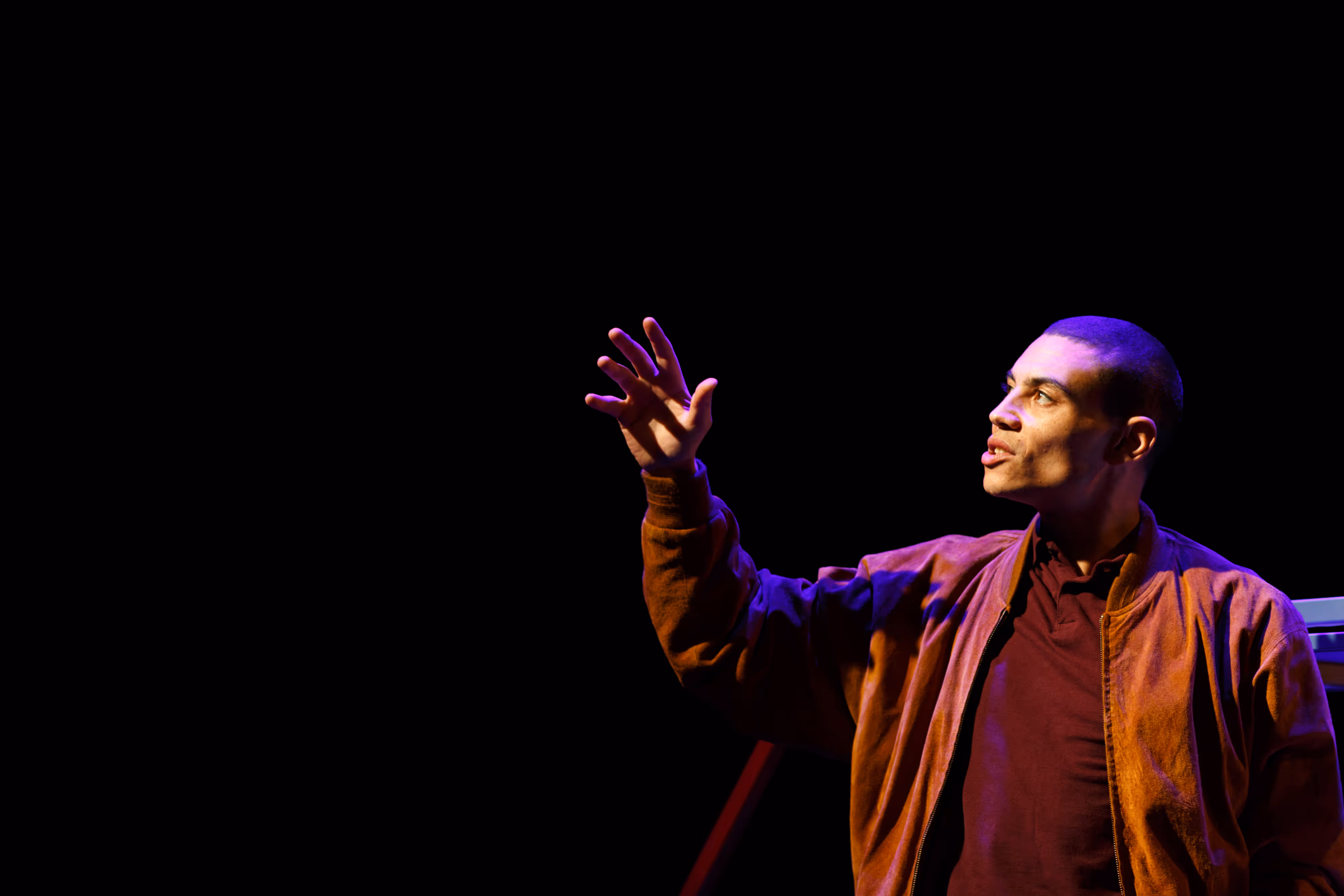A brown-jacketed performer gestures mid-speech, captured under moody stage lighting on a darkened theatre set.