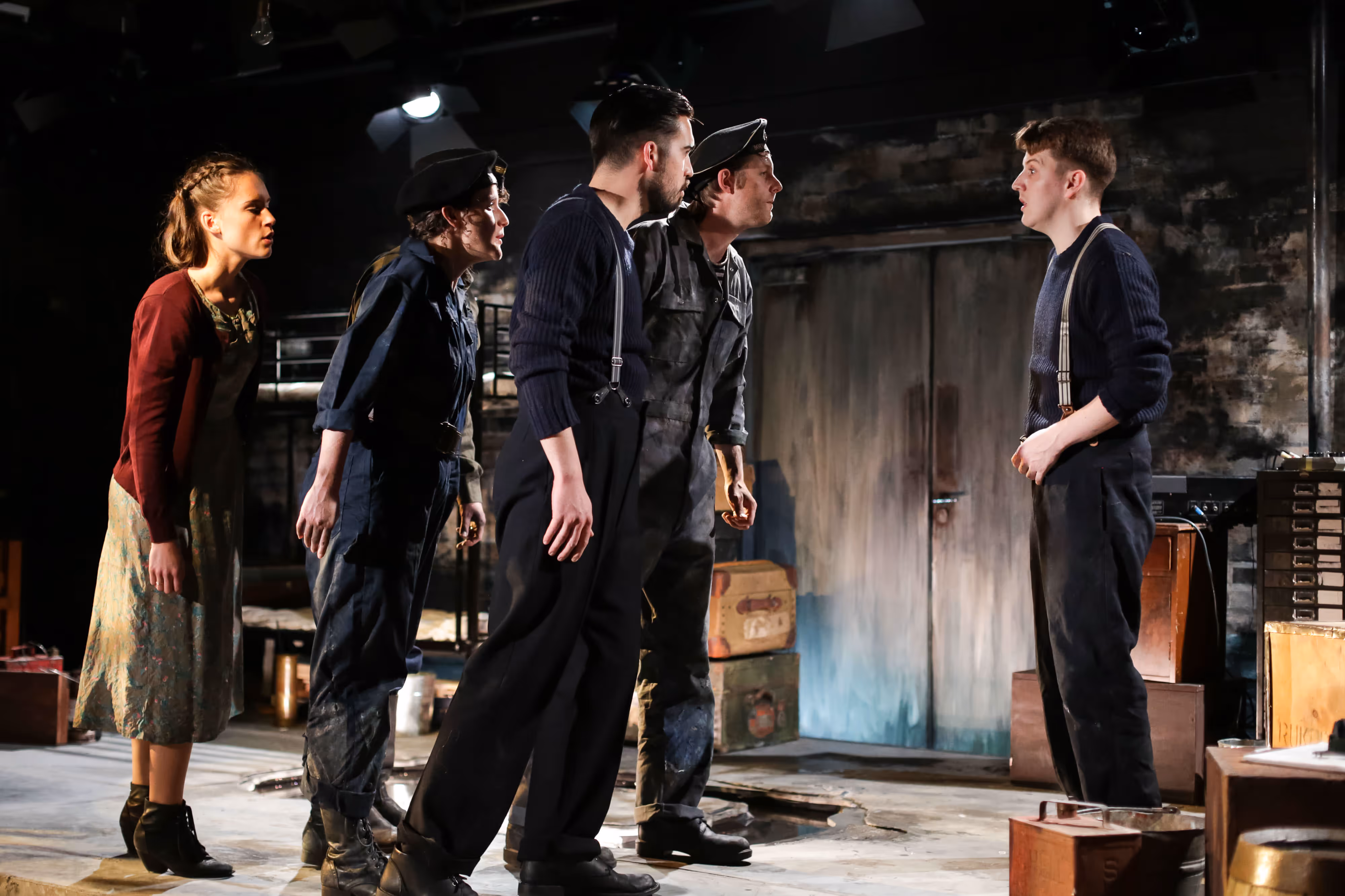 Under moody stage lighting, five actors in period costume pose dramatically, four aligned against one amid old crates and suitcases.