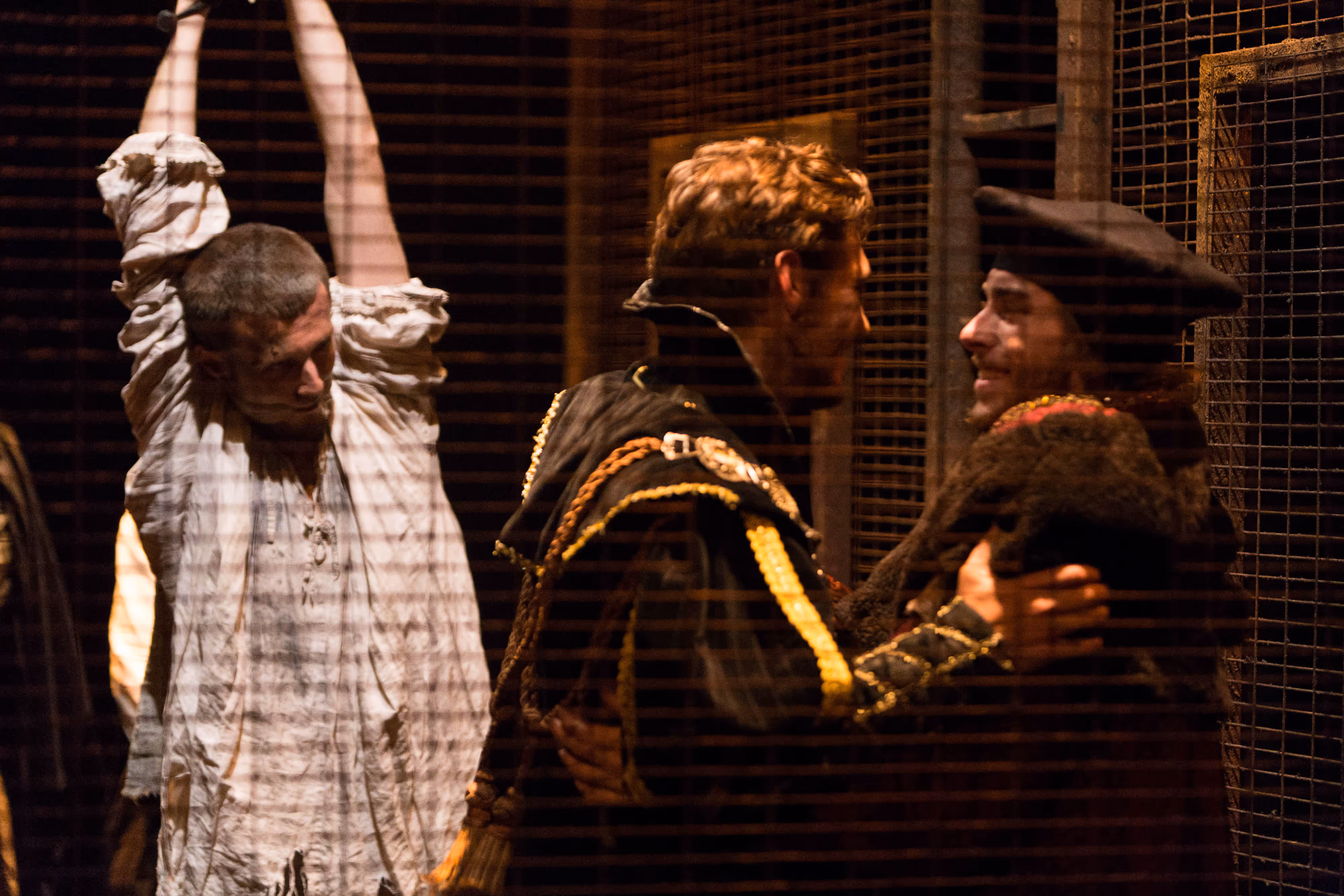 On stage, a chained actor raises his arms as two others in period costumes converse nearby behind a wire mesh.