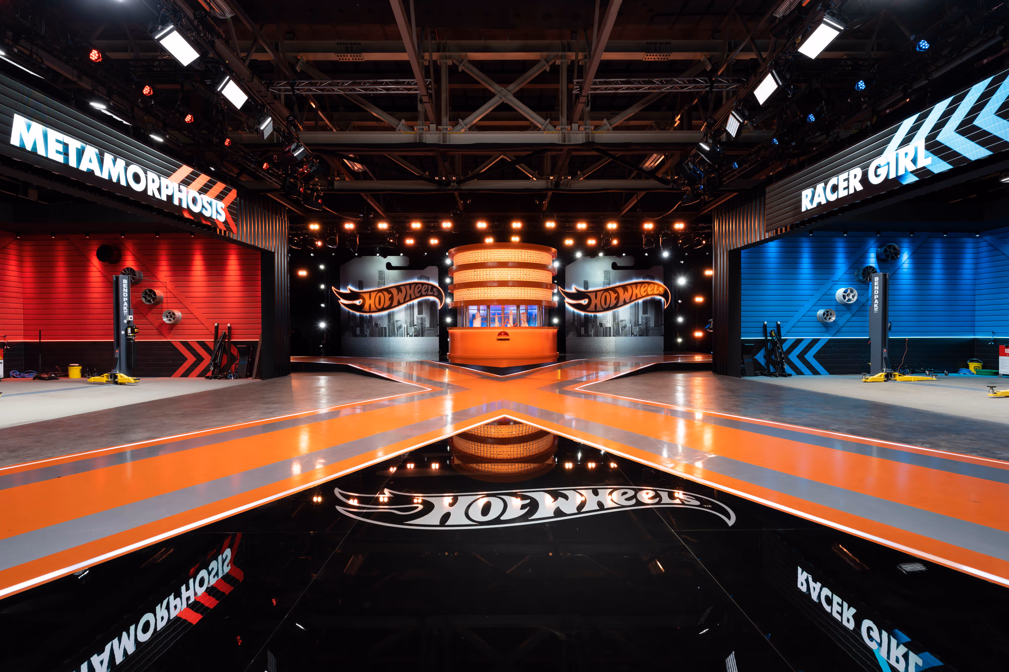 A vibrant tv set, illuminated like a theatre set, showcases Hot Wheels zones for "Metamorphosis" and "Racer Girl," red and blue-lit.