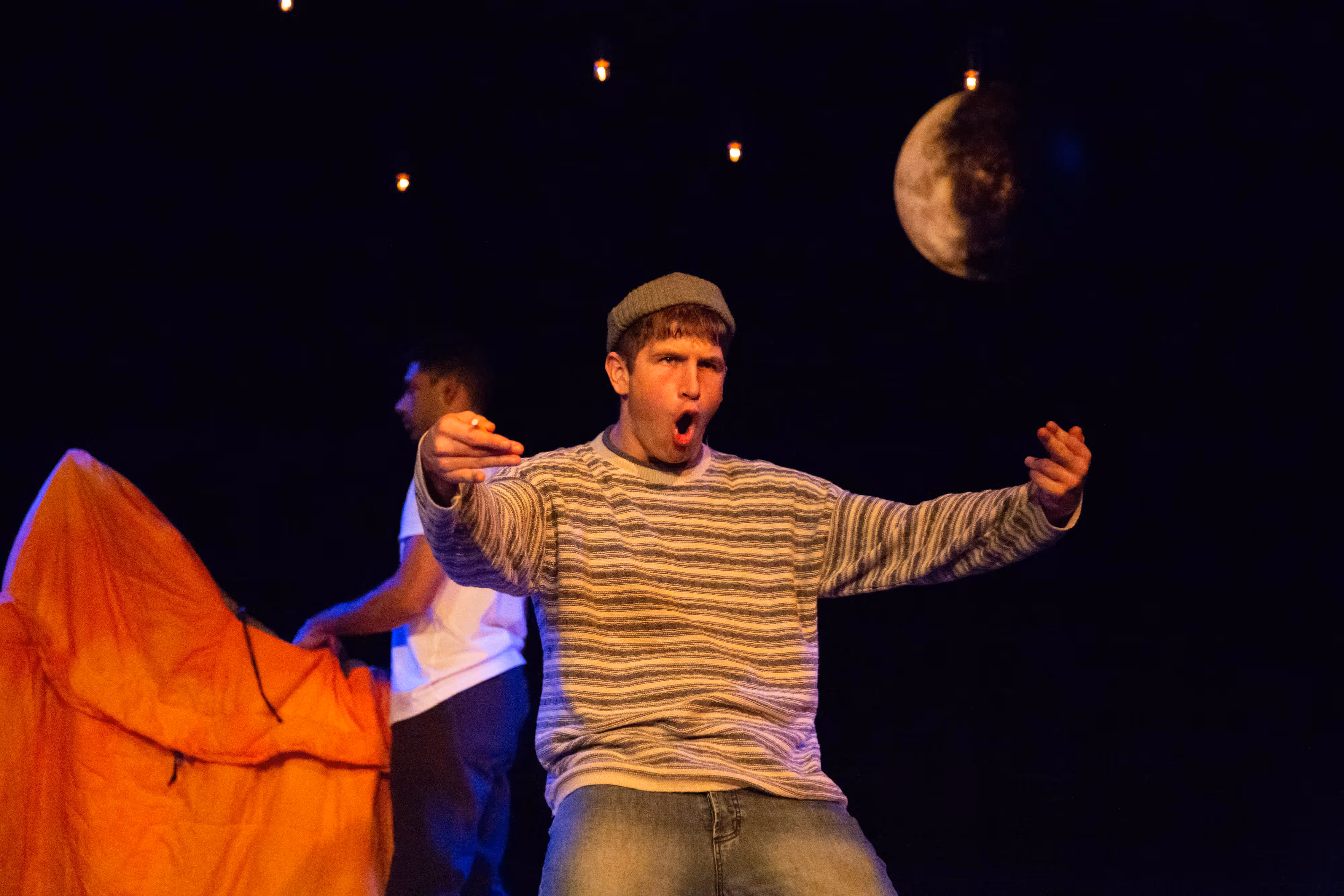 A performer in a striped jumper captured mid-gesture on stage, framed by a moon prop and glowing fairy lights behind him.
