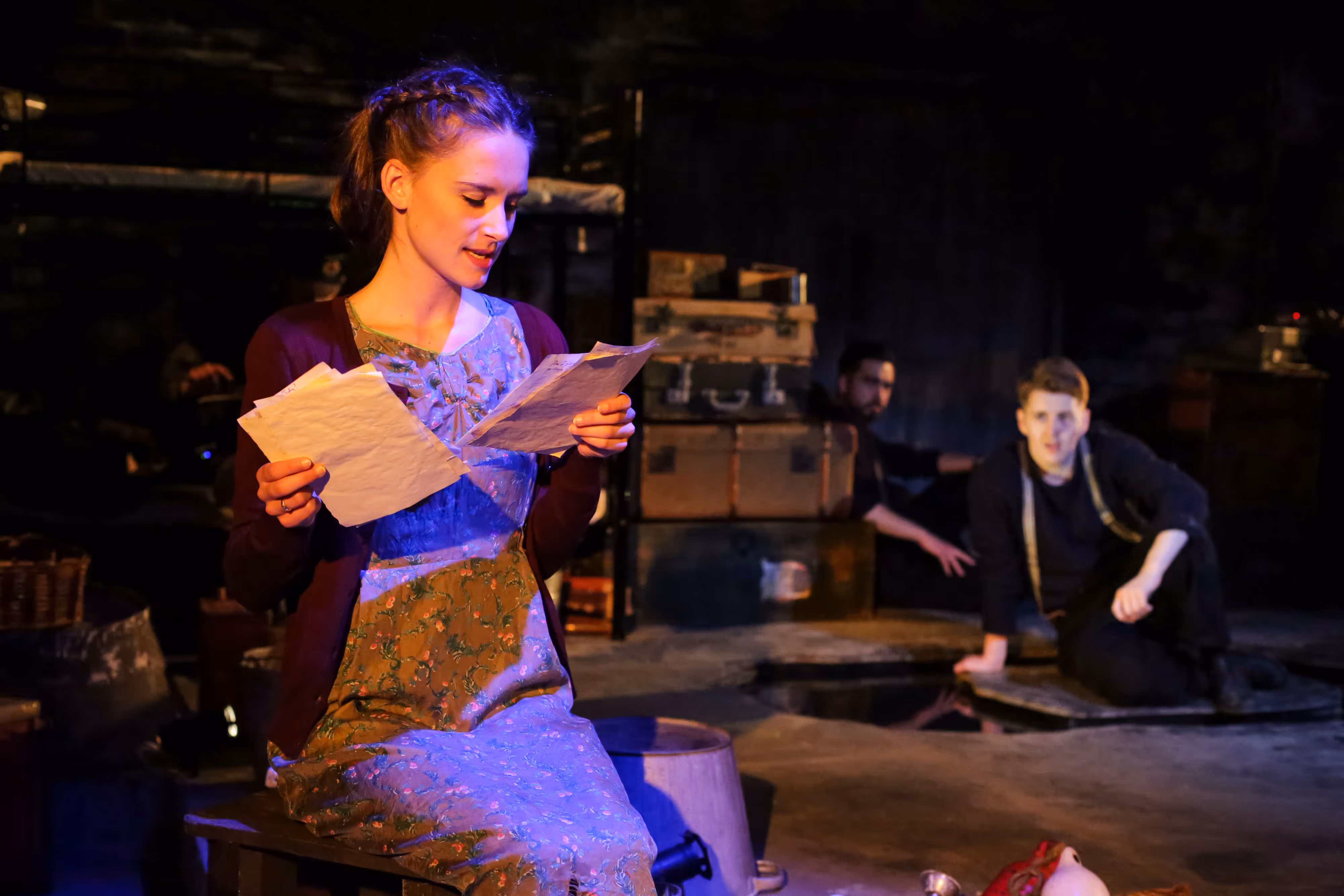 On stage, a woman reads a letter as two men sit behind her amid stacked suitcases, dramatic lighting casting deep shadows.