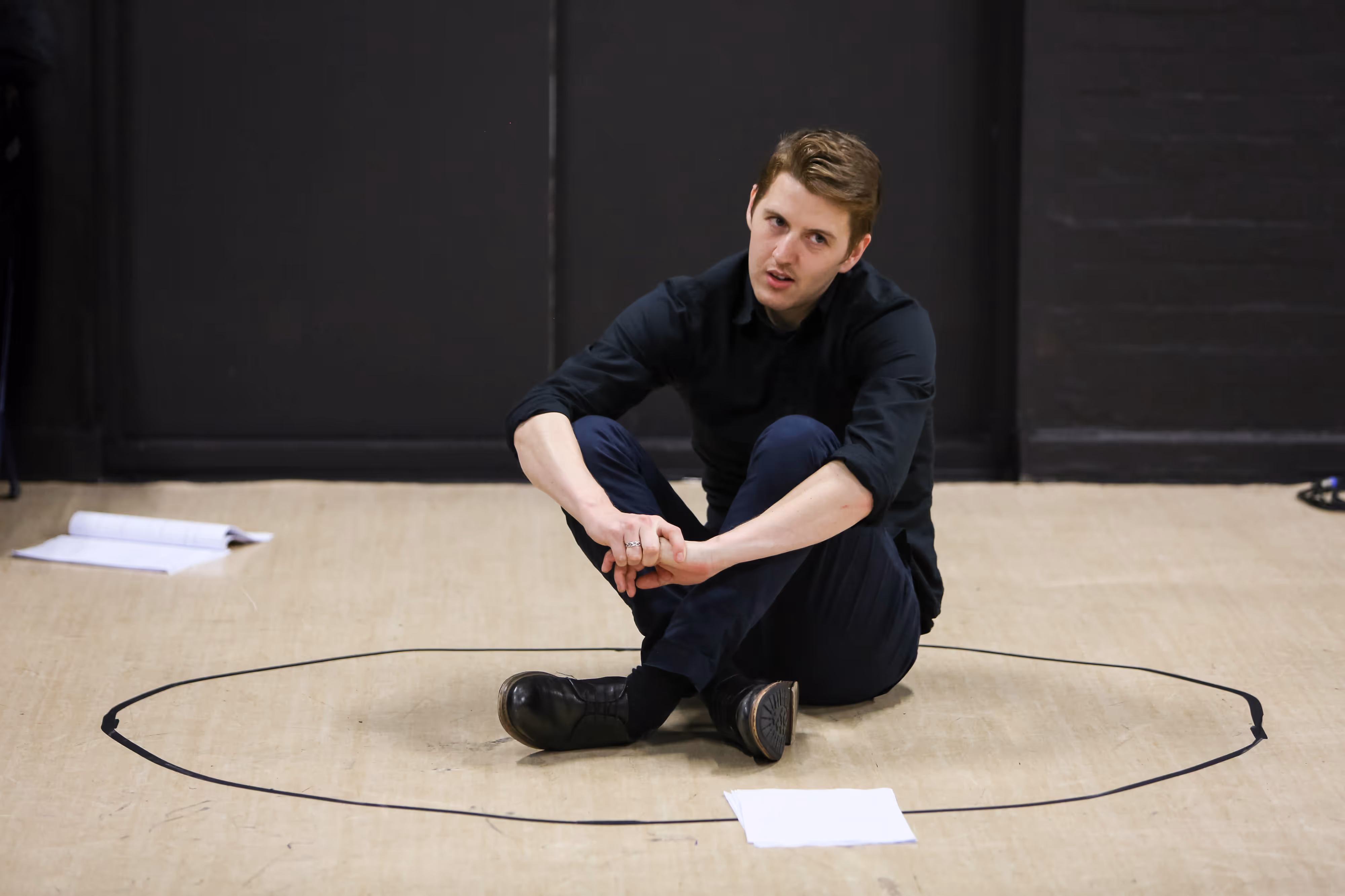 A performer in black sits cross-legged within a chalk circle centre stage, scattered scripts beside him, gazing upward into the lights.