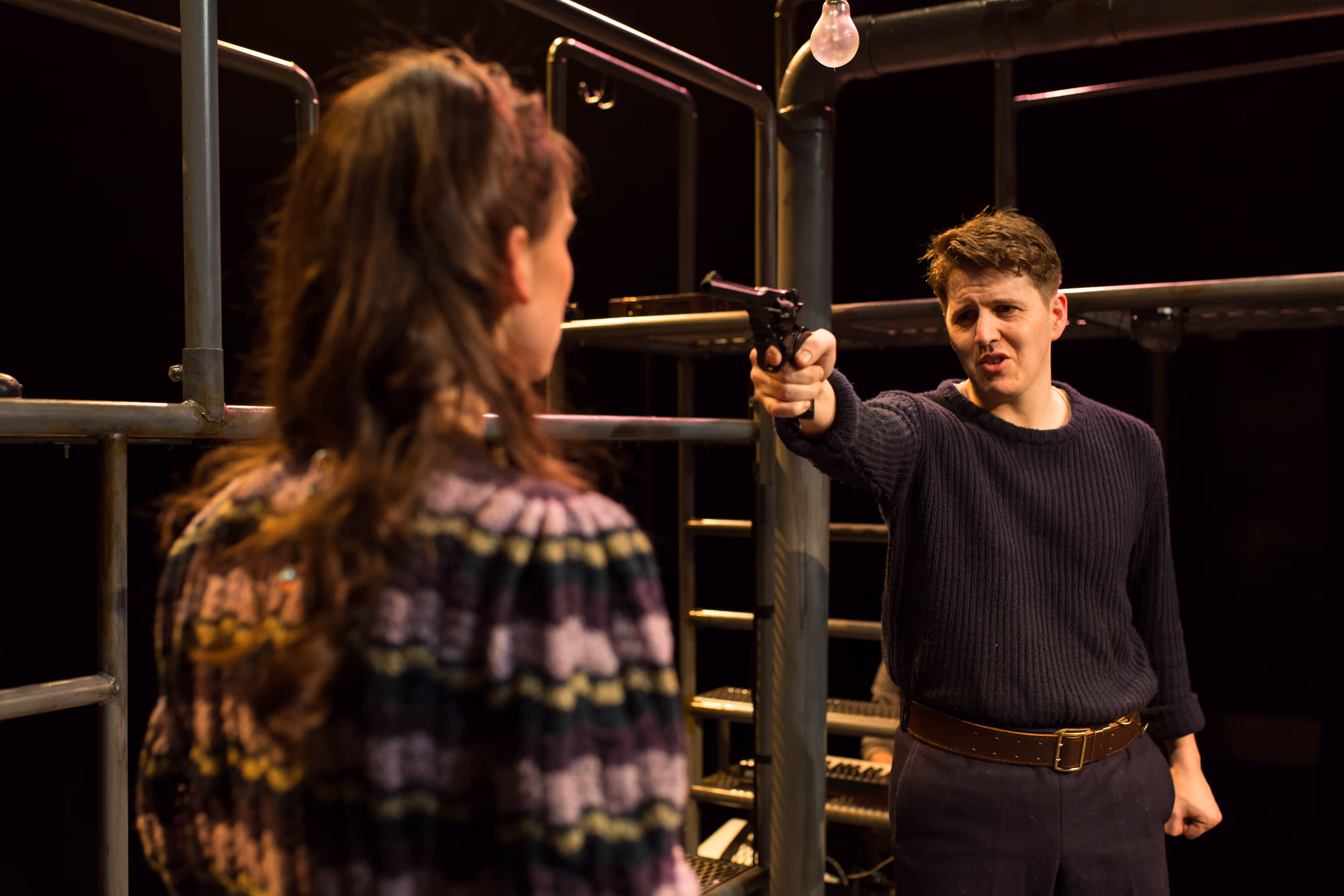 On stage, under dramatic lighting, an actor aims a prop gun at an actress on a shadowy metal staircase, both in intense poses.