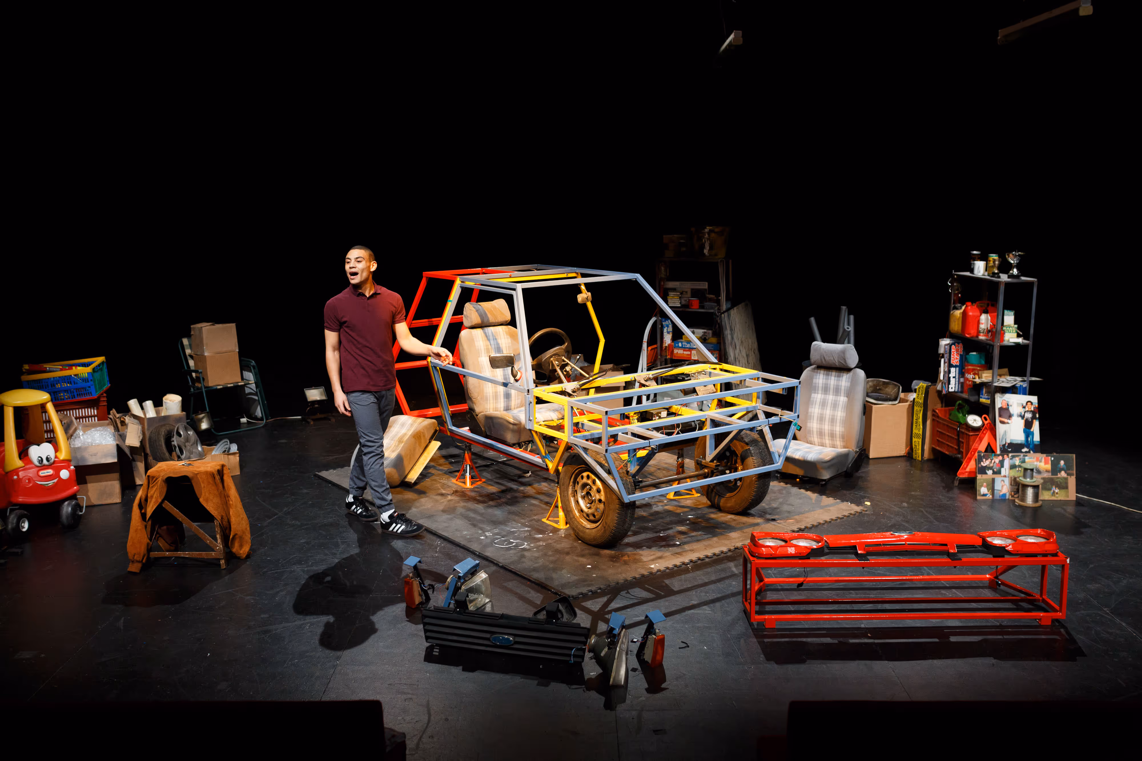 A theatre photographer captures a person beside a vibrant car body on stage, amid scattered parts and boxes in a set design.