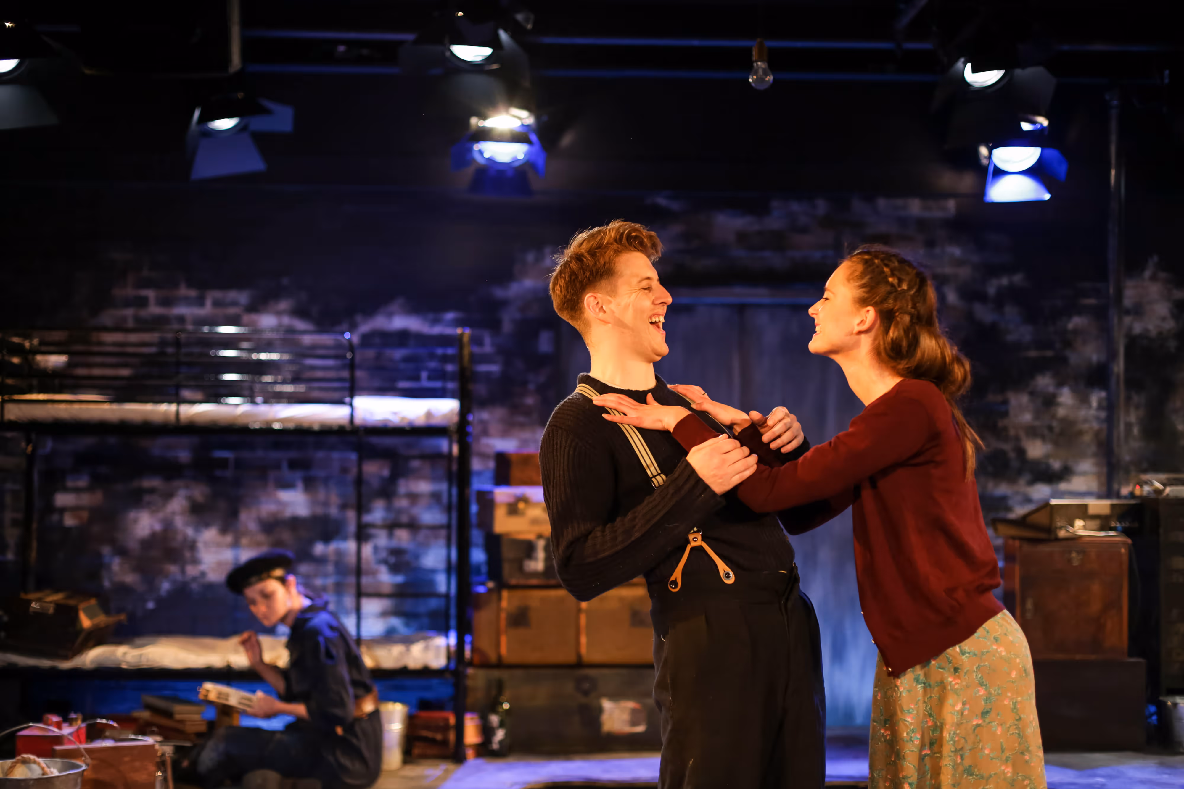 From the wings: two cast members beam, holding hands centre stage, as another sits by bunk beds and luggage upstage.