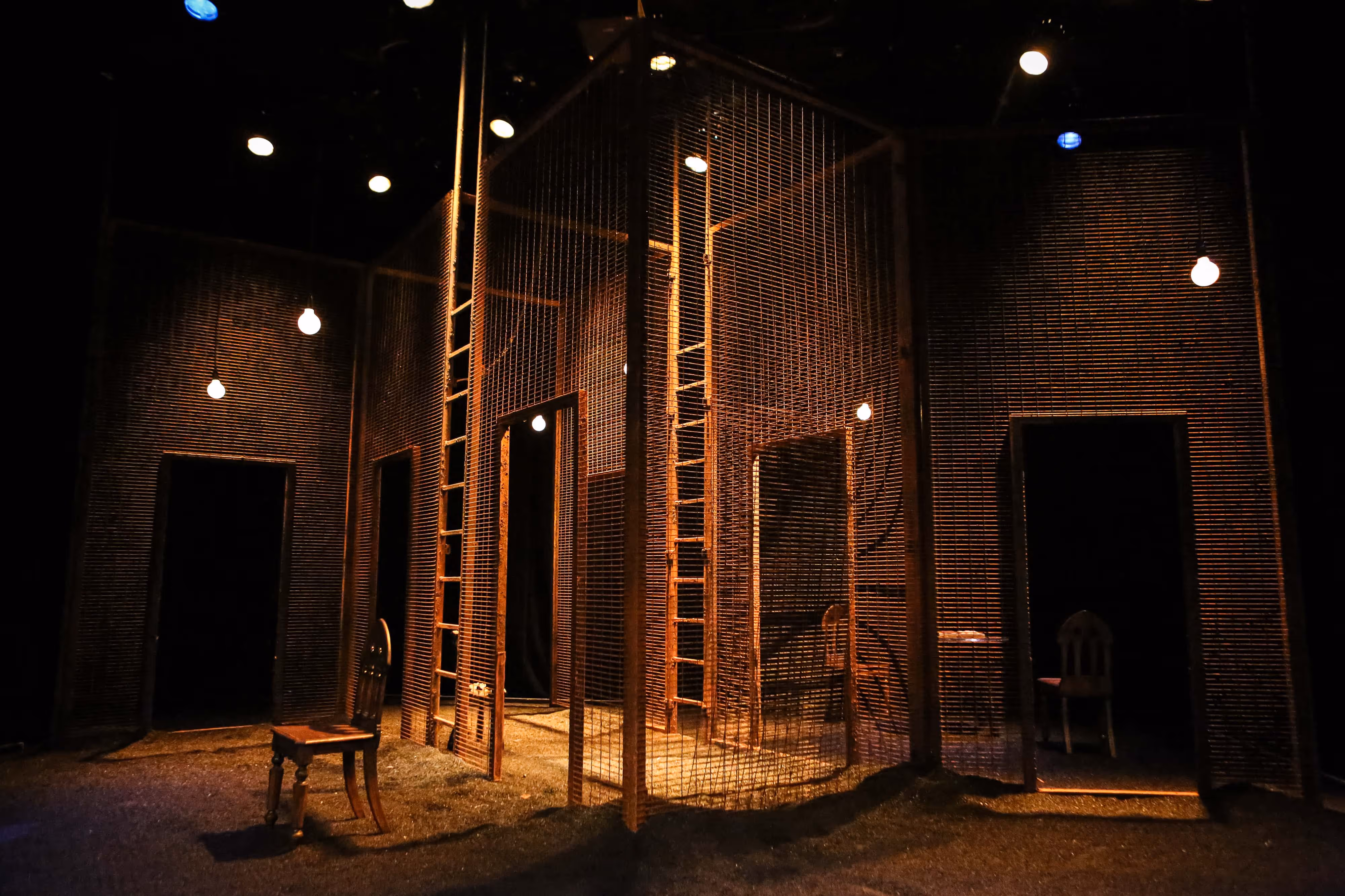 Moody stage scene: ladders, open door frames, suspended bulbs, and wooden chairs arranged in a wire-frame house set for theatre.