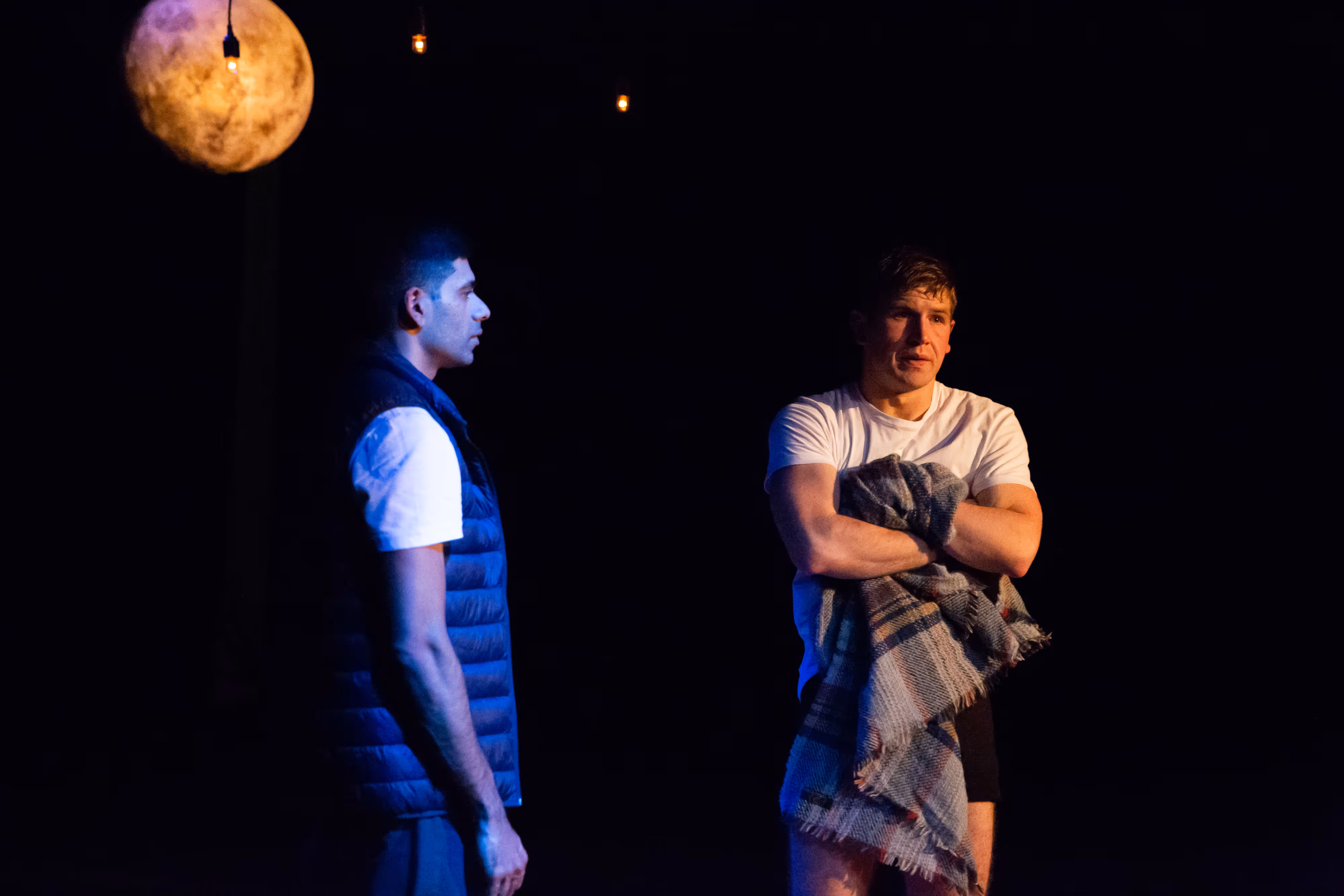 On stage, two men under moody lighting; one draped in a blanket, both maintain intense expressions as they face each other.