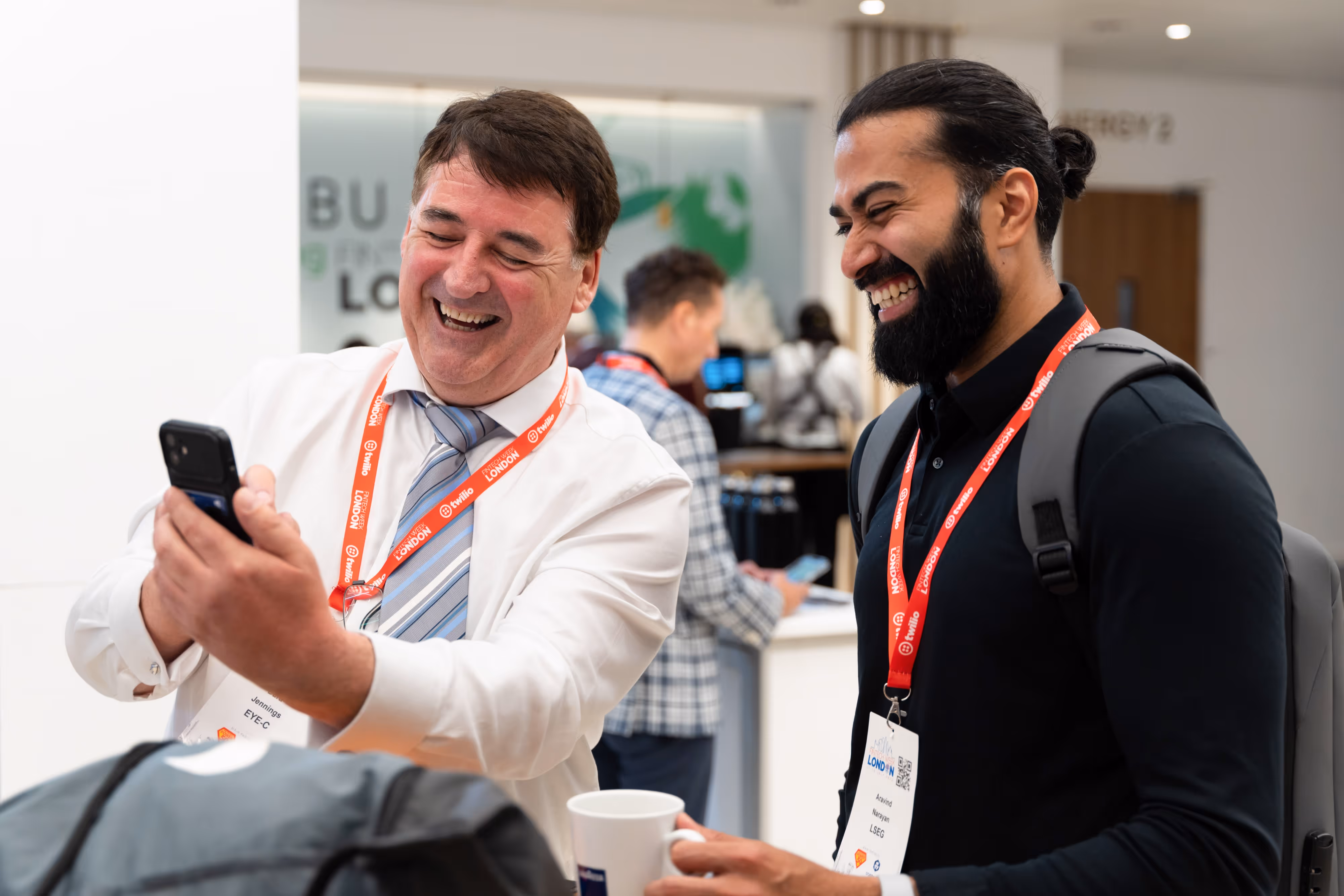 Two men wearing conference badges smile and laugh whilst taking a selfie together at an indoor event.