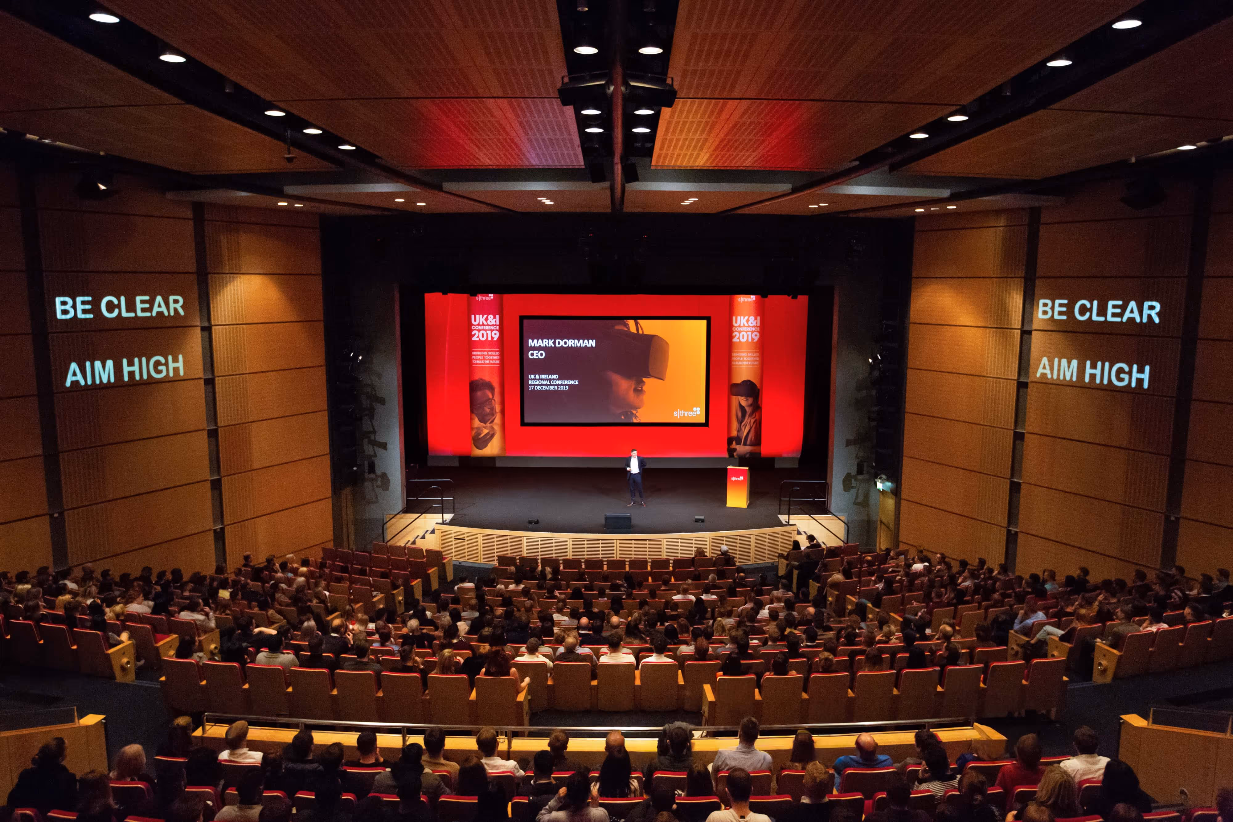 A speaker presents on stage to a full auditorium; screens display the phrases "BE CLEAR" and "AIM HIGH".