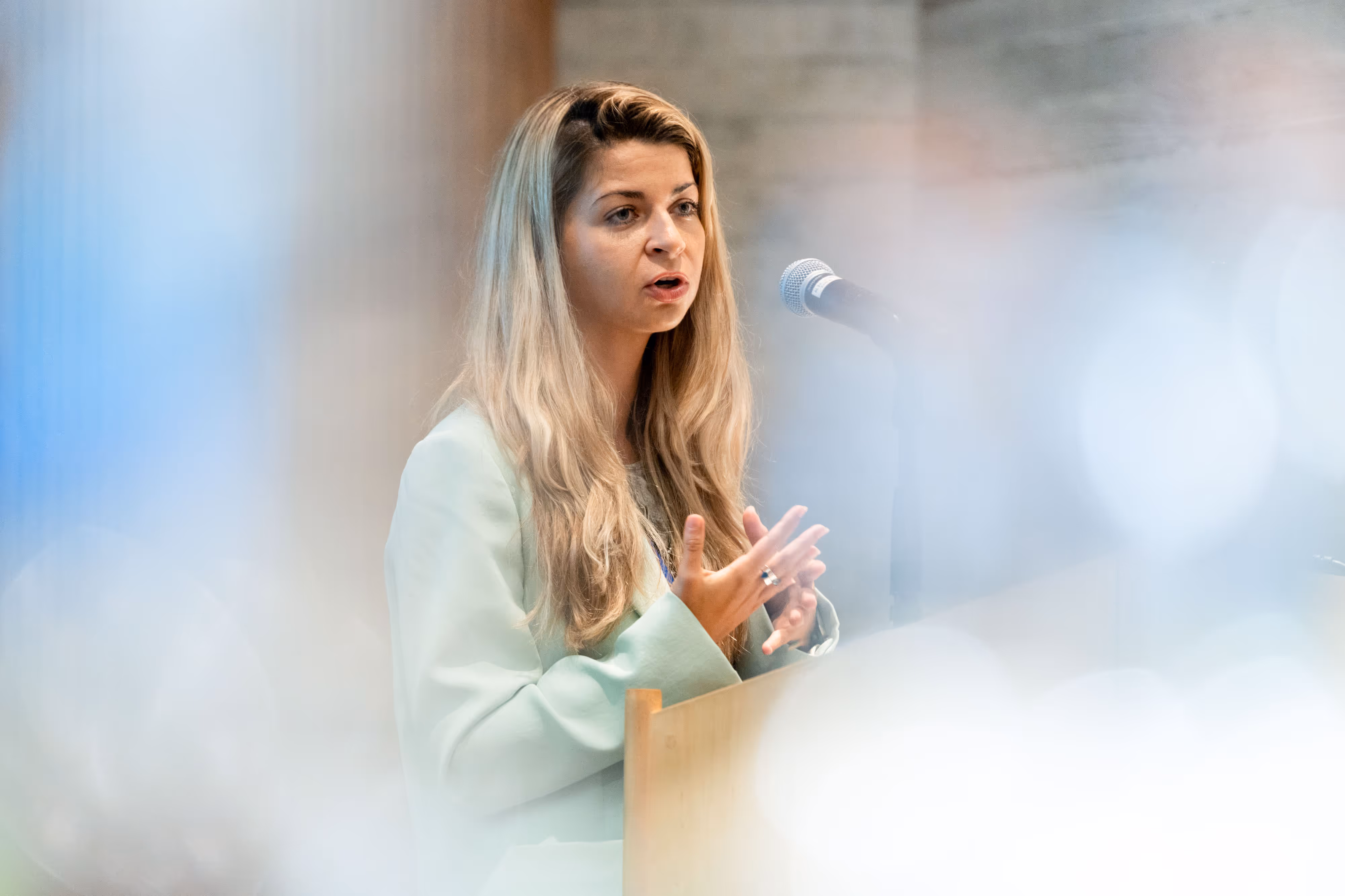 Woman with long blonde hair speaking at a lectern with a microphone, blurred lights in the foreground.