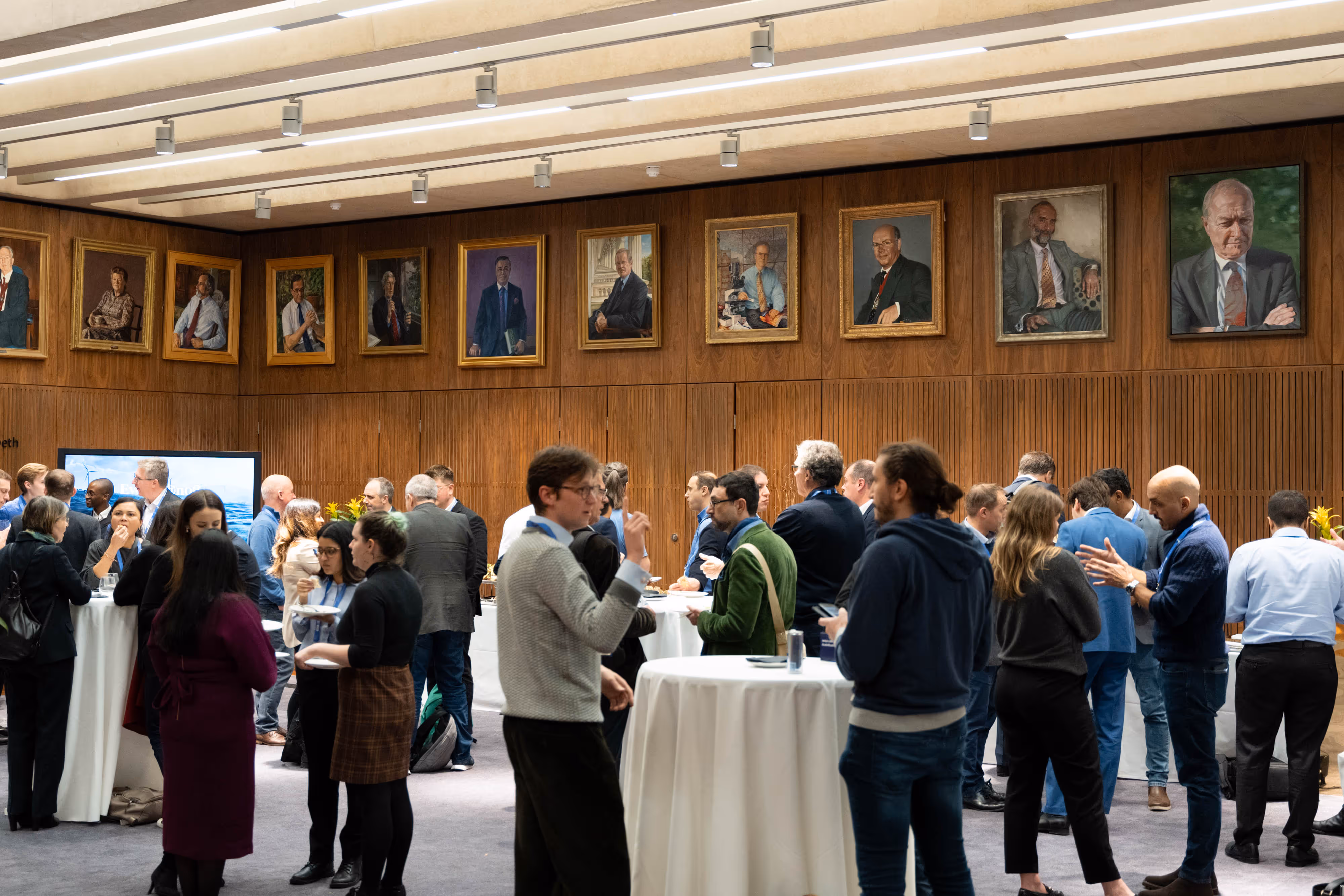 People networking at tall tables in a large room with portraits of distinguished individuals on wood-panelled walls.