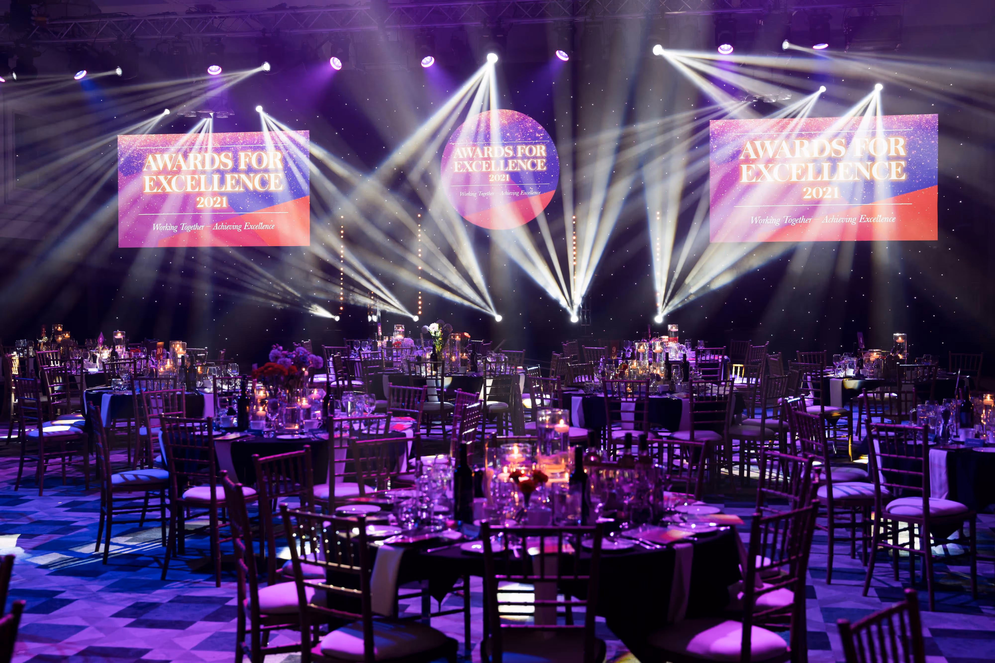 A decorated ballroom with round tables, stage lights, and screens reading "Awards for Excellence 2021".