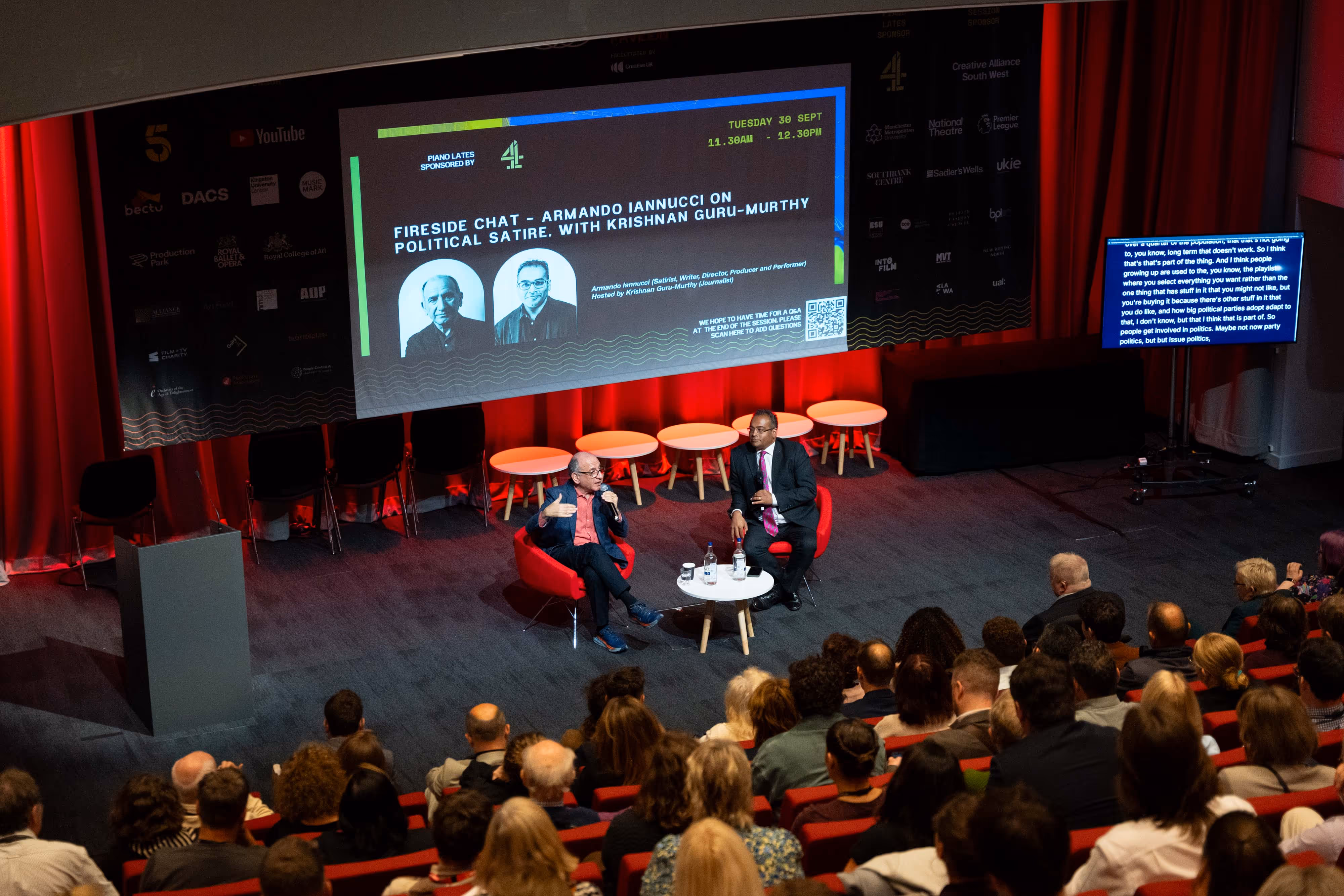 Two men sit on stage in armchairs, speaking to an audience, with a large event screen and red curtains behind them.