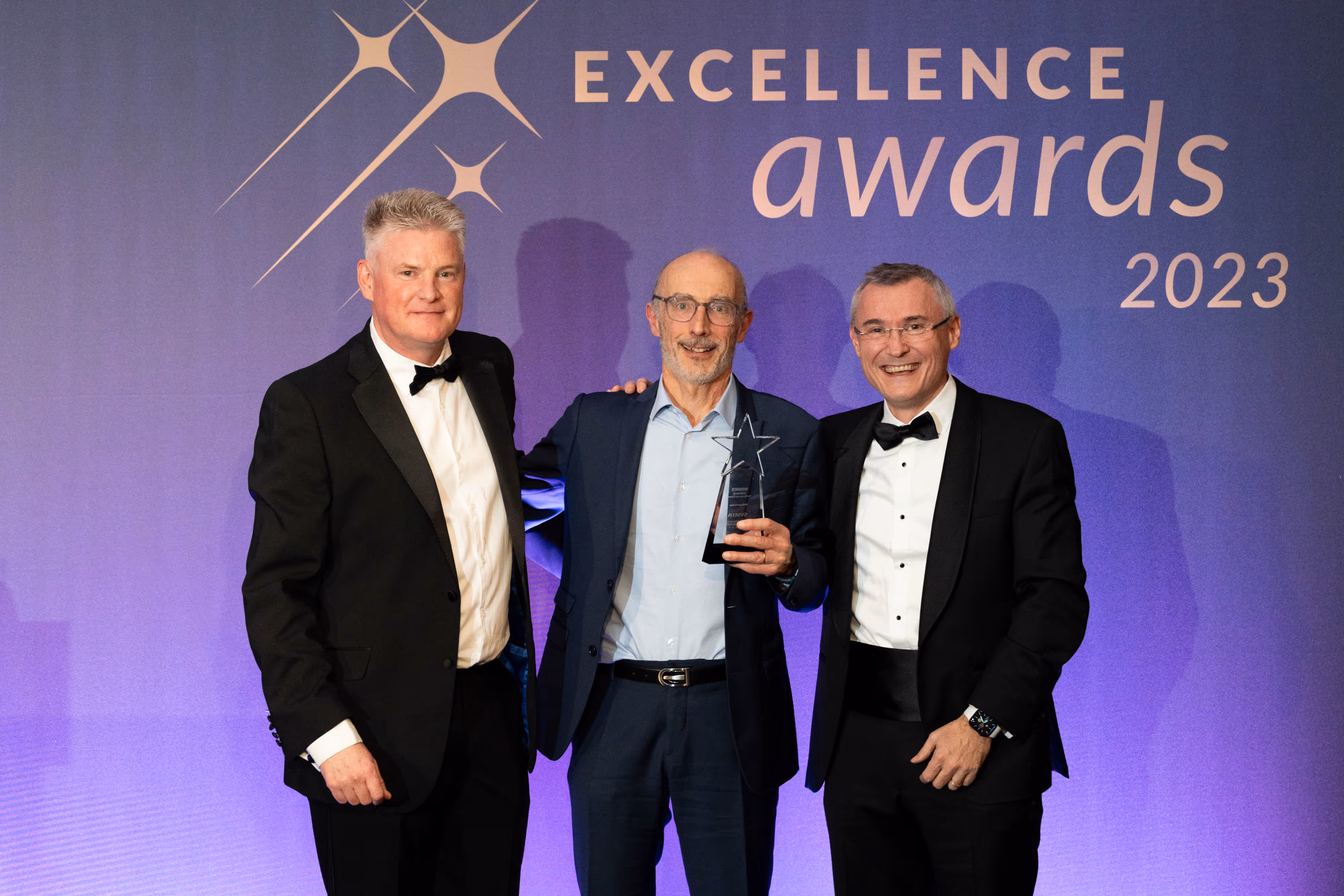 Three men in suits and tuxedos pose with an award at the 2023 Excellence Awards against a purple backdrop.