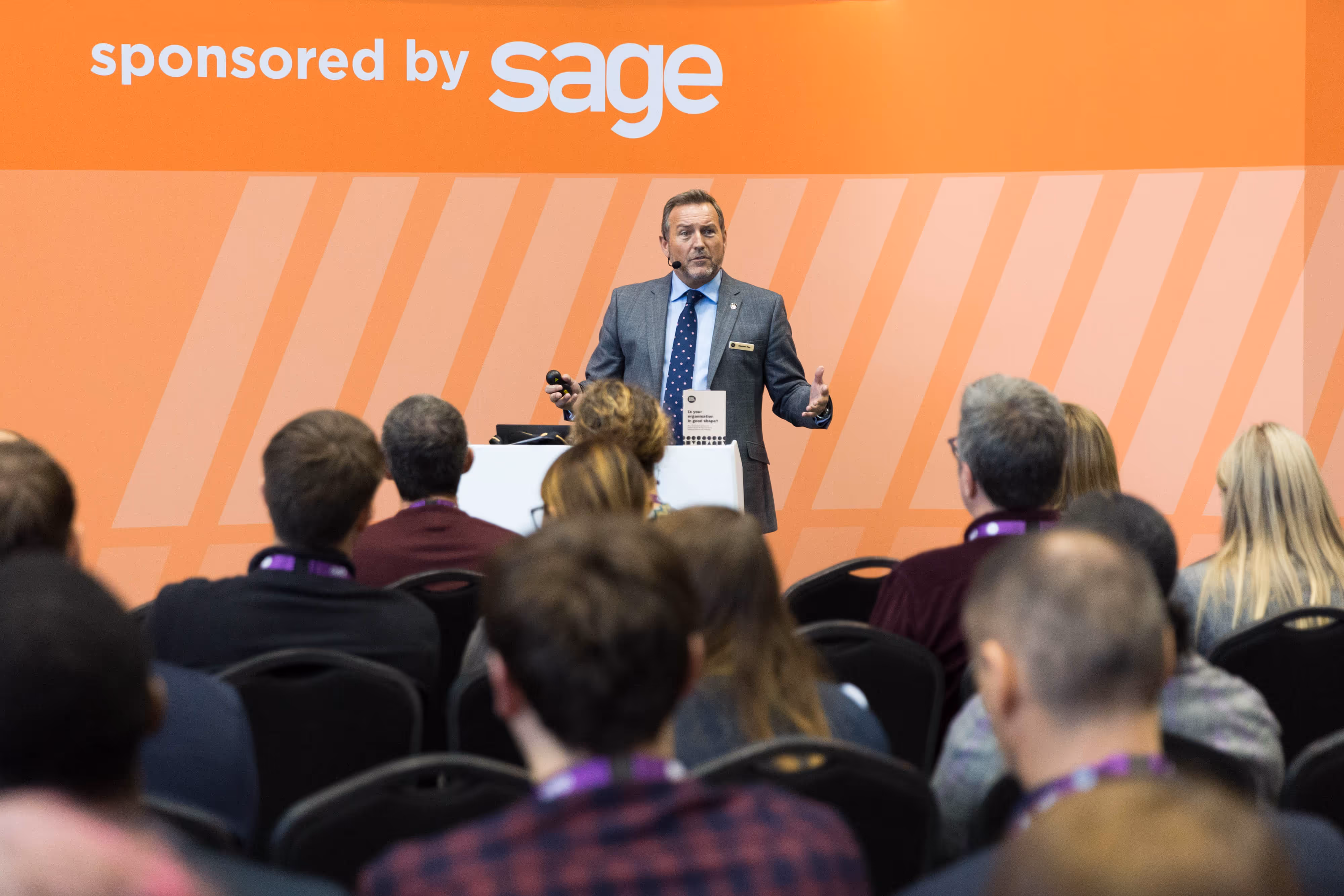 A man speaks on stage to an audience at a conference, in front of an orange "sponsored by sage" backdrop.