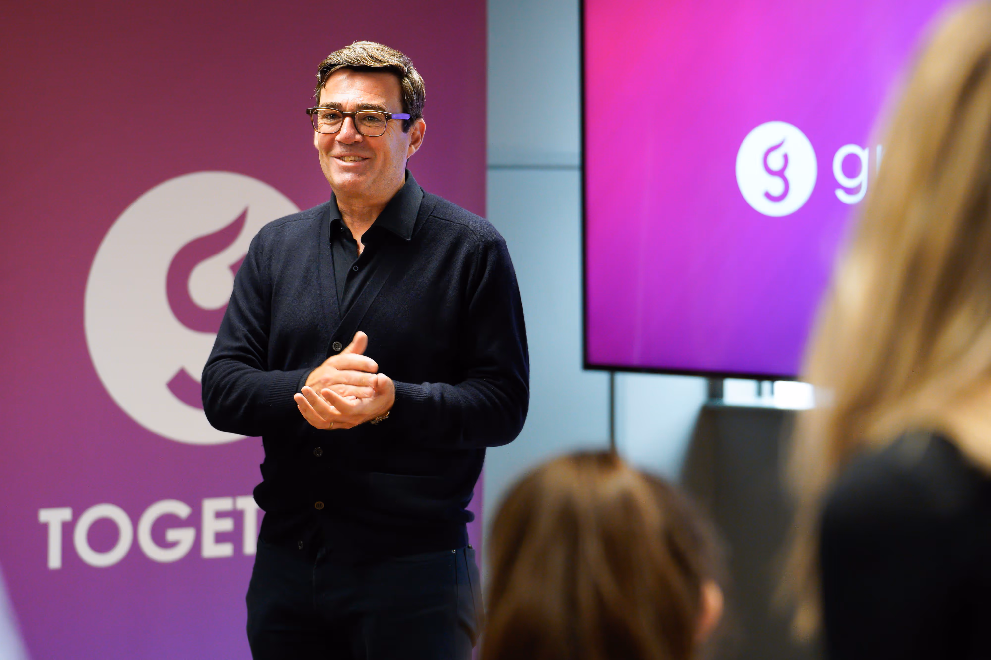 Mayor of Greater Manchester Andy Burnham speaks and smiles in front of a purple "Together" sign to an audience.