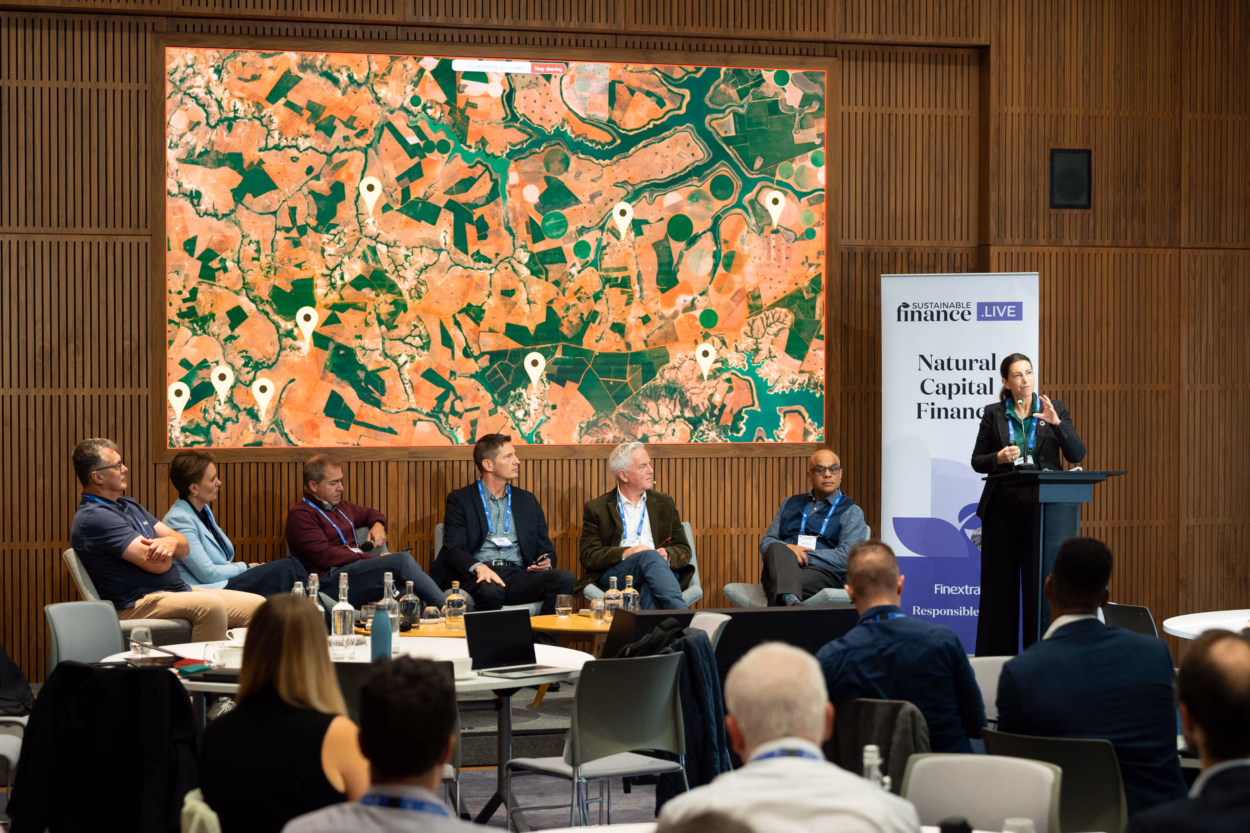 A panel of six people sits onstage at a conference, with a speaker at a lectern and a colourful map displayed behind them.