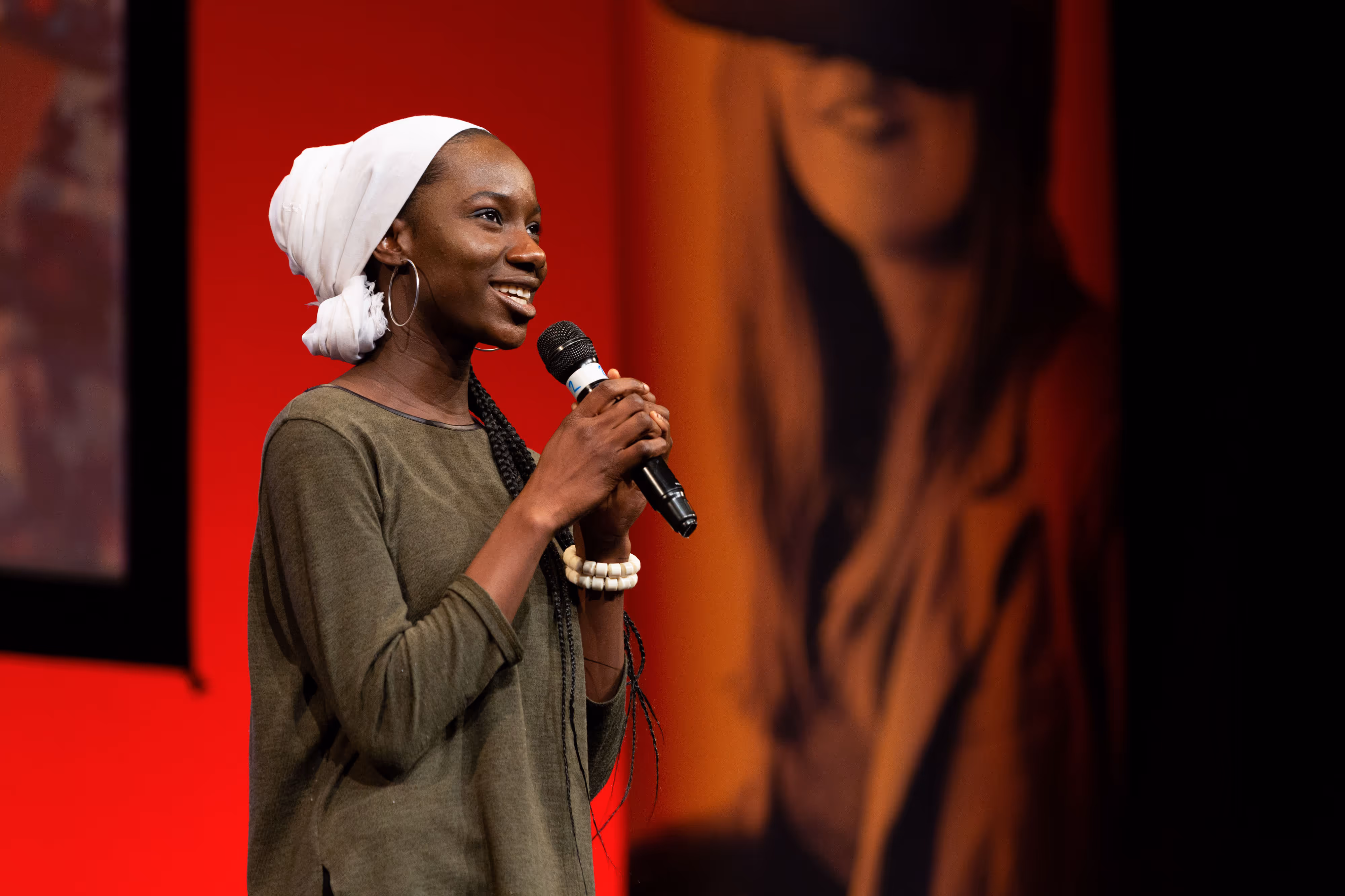 A woman in a green top and white headscarf speaks into a microphone on stage with a red and black background.