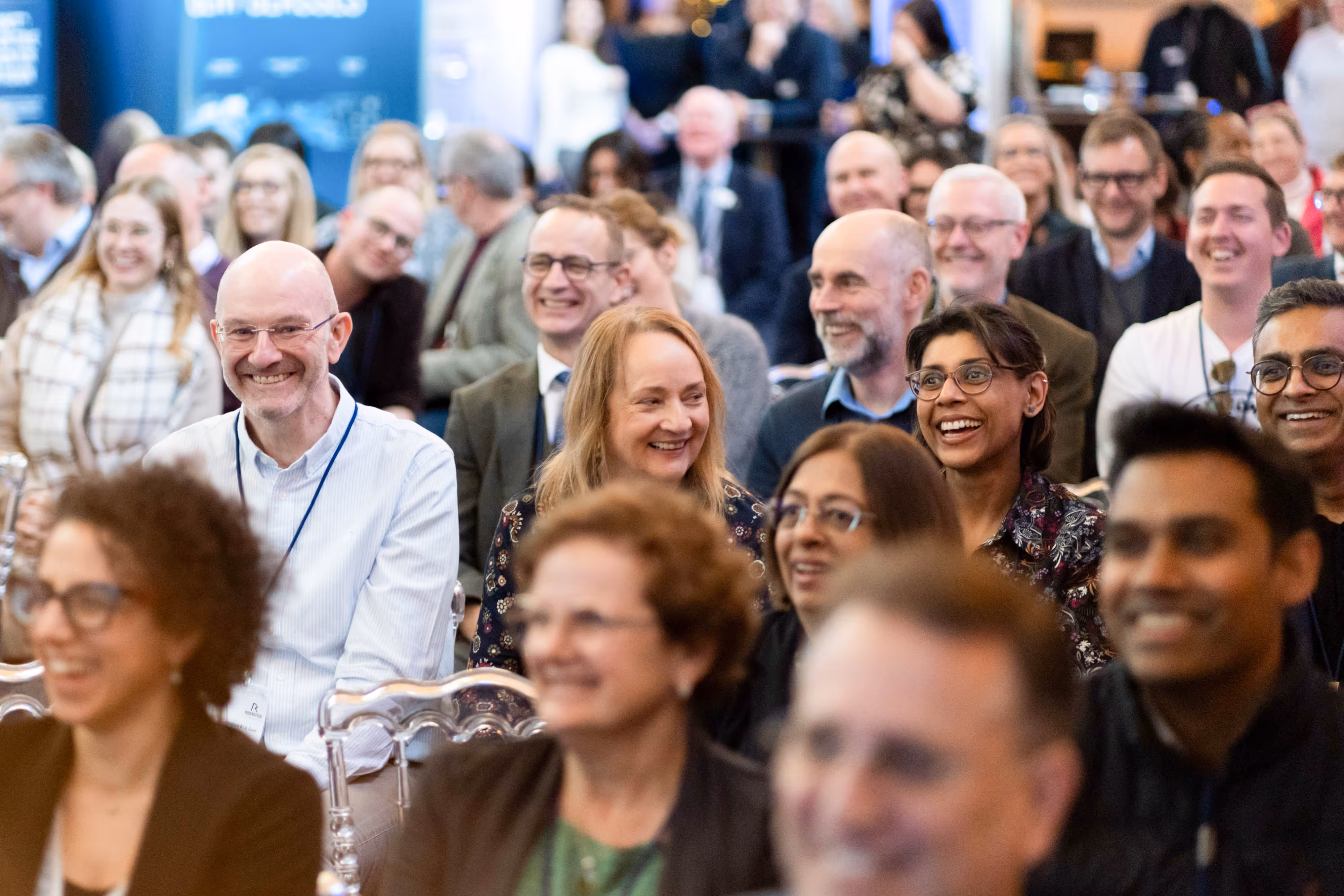 A diverse group of people sit closely together, smiling and laughing at an indoor event or conference.