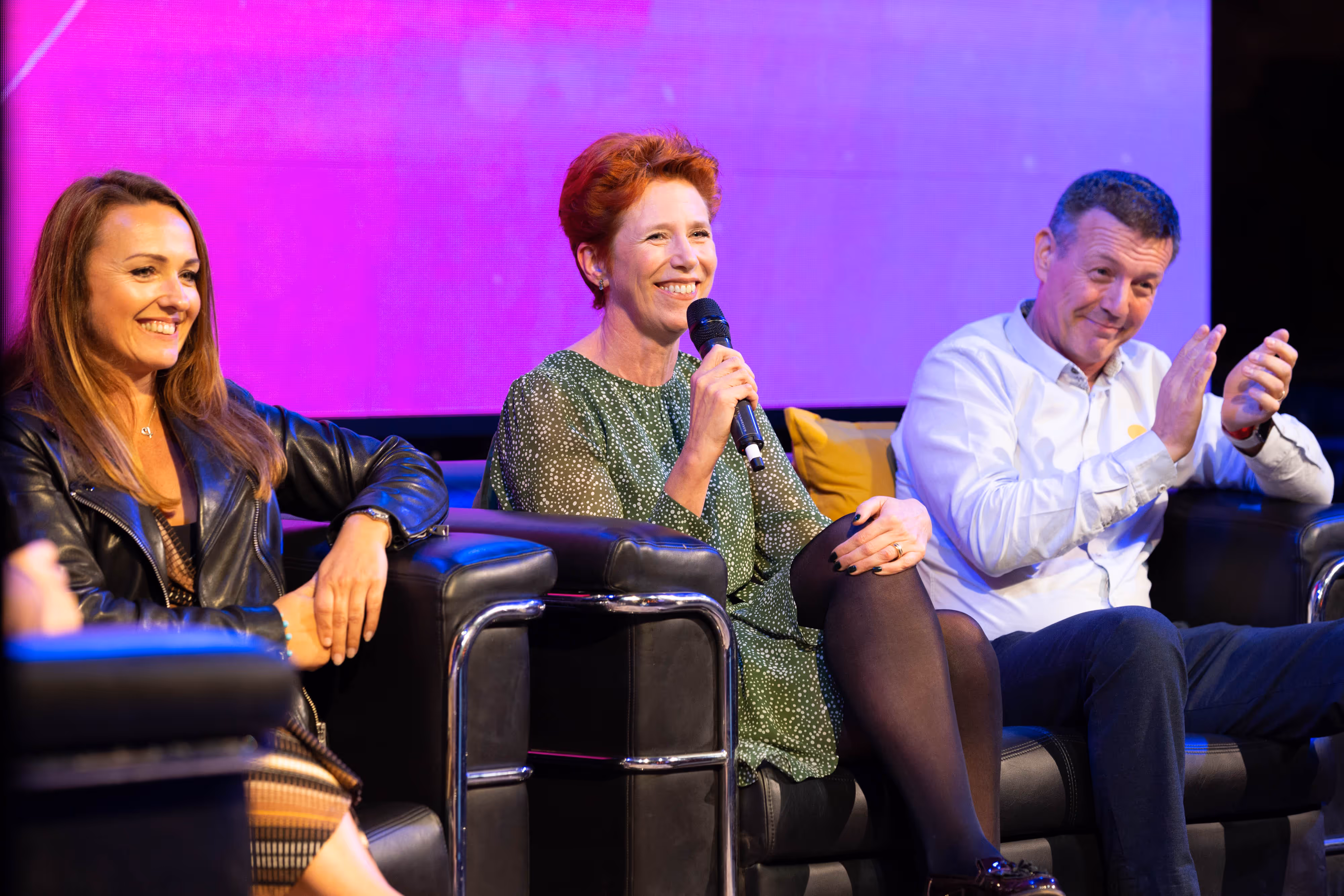 Three people sit on stage in chairs; the woman in the centre speaks into a microphone while the other two smile and clap.